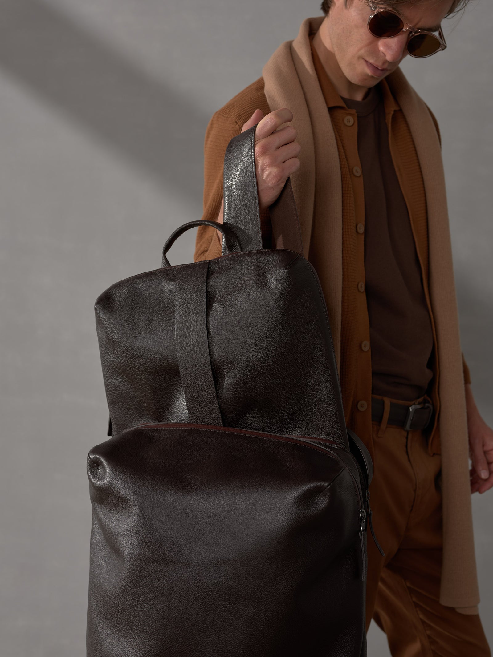 Man holding a luxury dark brown leather backpack by the top handle against a gray minimalist background.