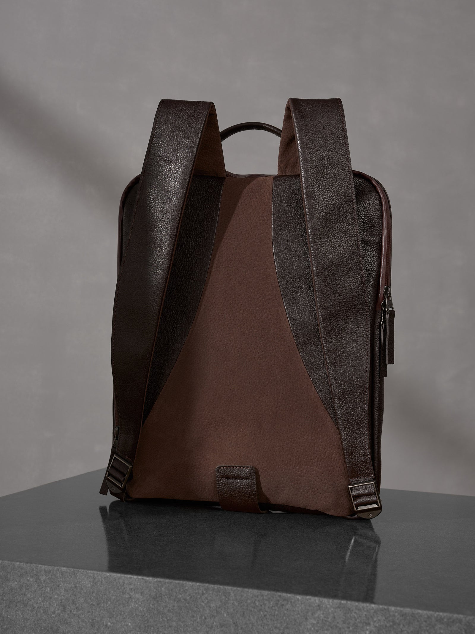 Rear view of a luxury dark brown Capra Leather backpack with ergonomic shoulder straps on a polished surface.