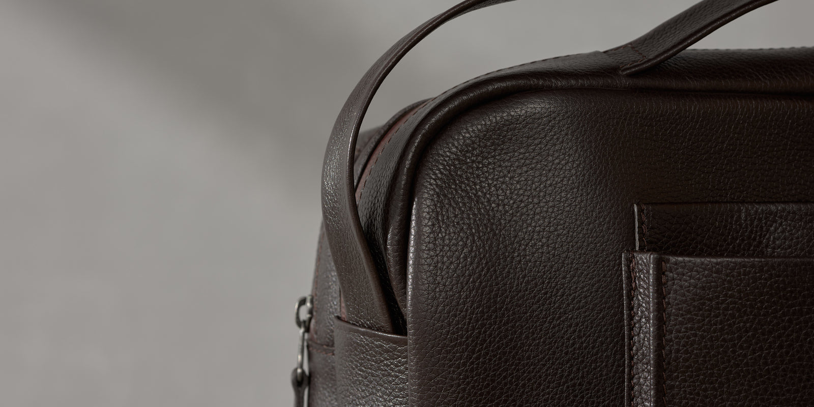 Macro shot of pebble-grain dark brown leather texture, side strap, and fine stitching on a premium bag.