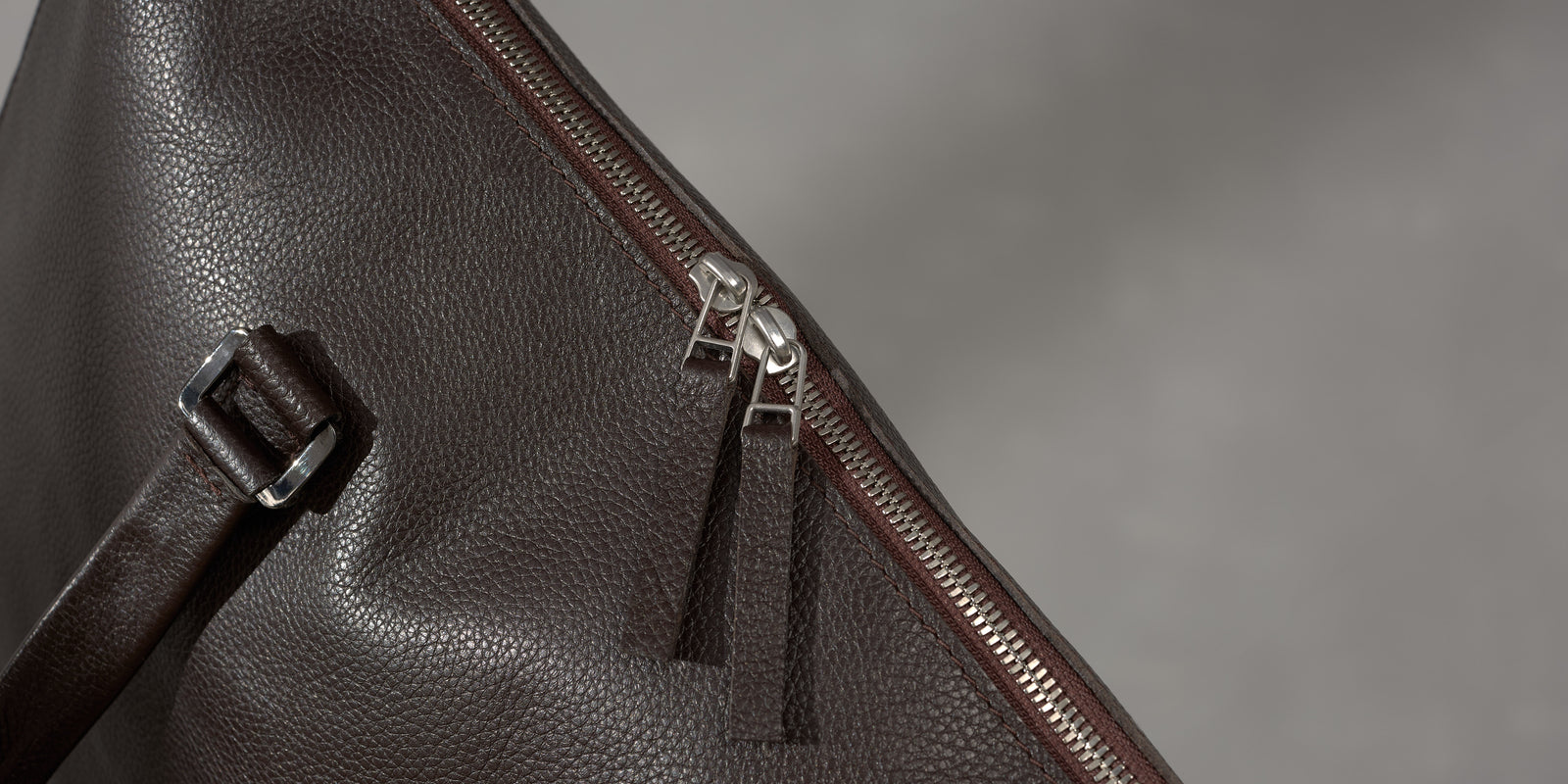 Extreme close-up of a silver metal dual zipper mechanism on a textured dark brown leather travel bag.