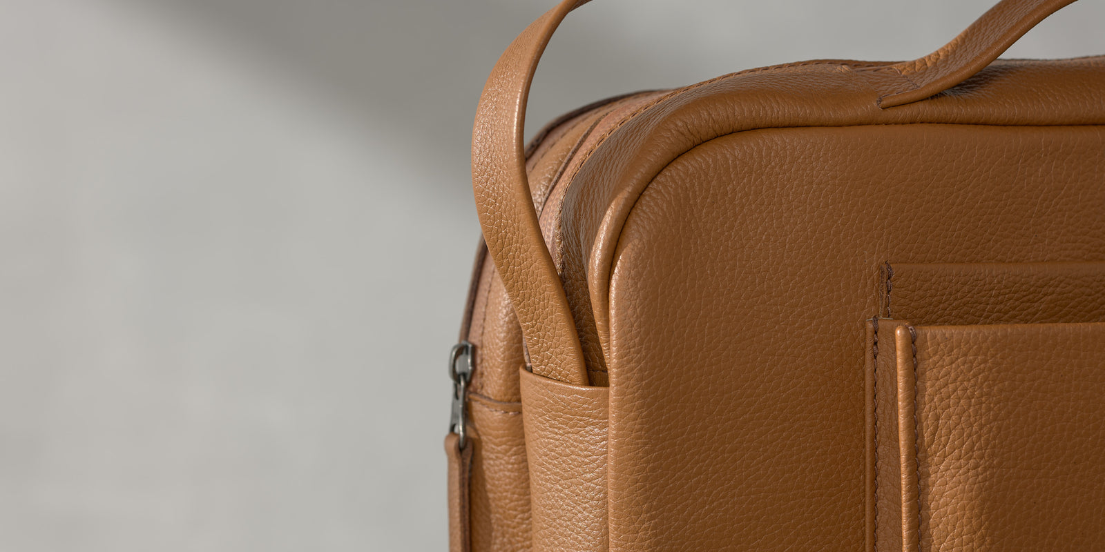Close-up of the textured tan leather, stitched pocket edge, and side handle attachment loop on a Capra Leather bag.