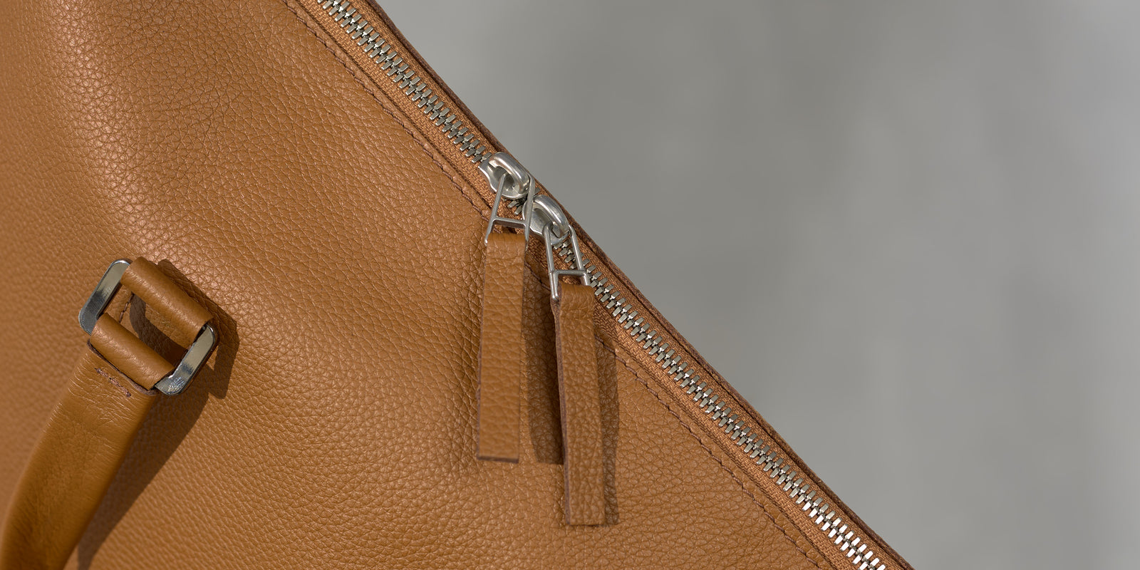 Close-up of a silver metallic zipper with two tan leather pull tabs on a full-grain leather bag.