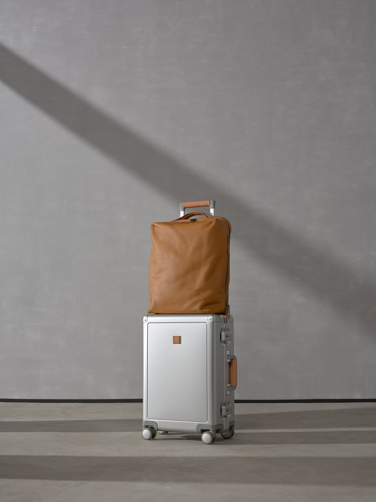 Tan full-grain leather carry-on bag from the Basalt System sitting on top of a sleek silver aluminum rolling suitcase against a gray background.