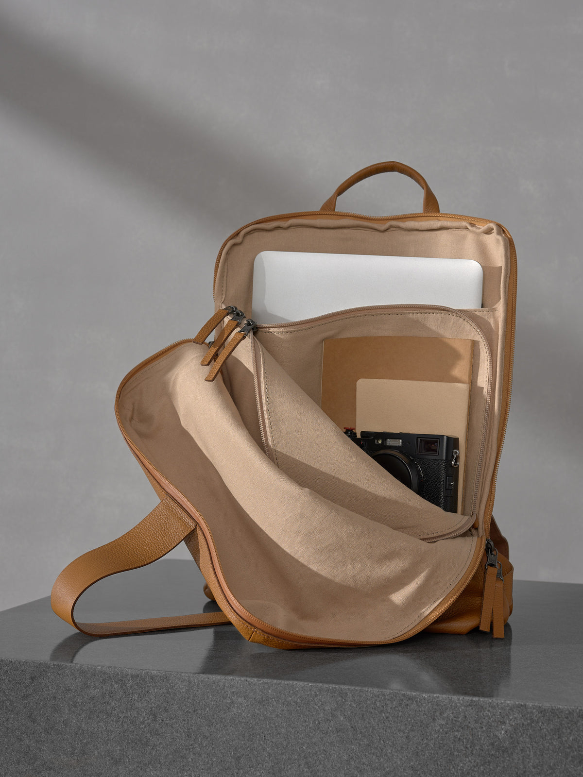 Open interior view of the Capra Leather tan backpack displaying its organized compartments containing a laptop, notebooks, and a black camera.