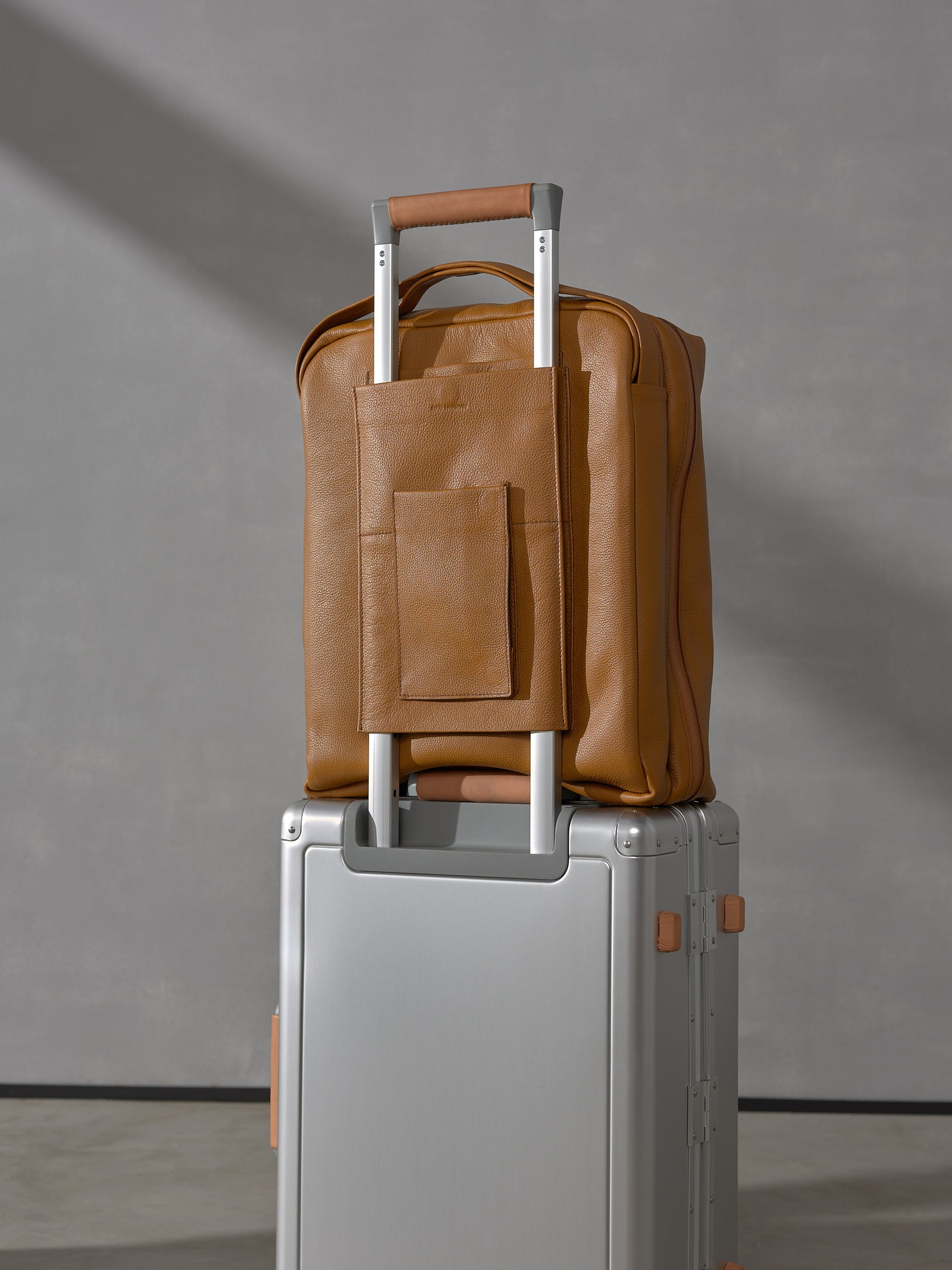 Back view of the tan leather carry-on bag attached to a silver suitcase telescopic handle via its integrated leather trolley sleeve.