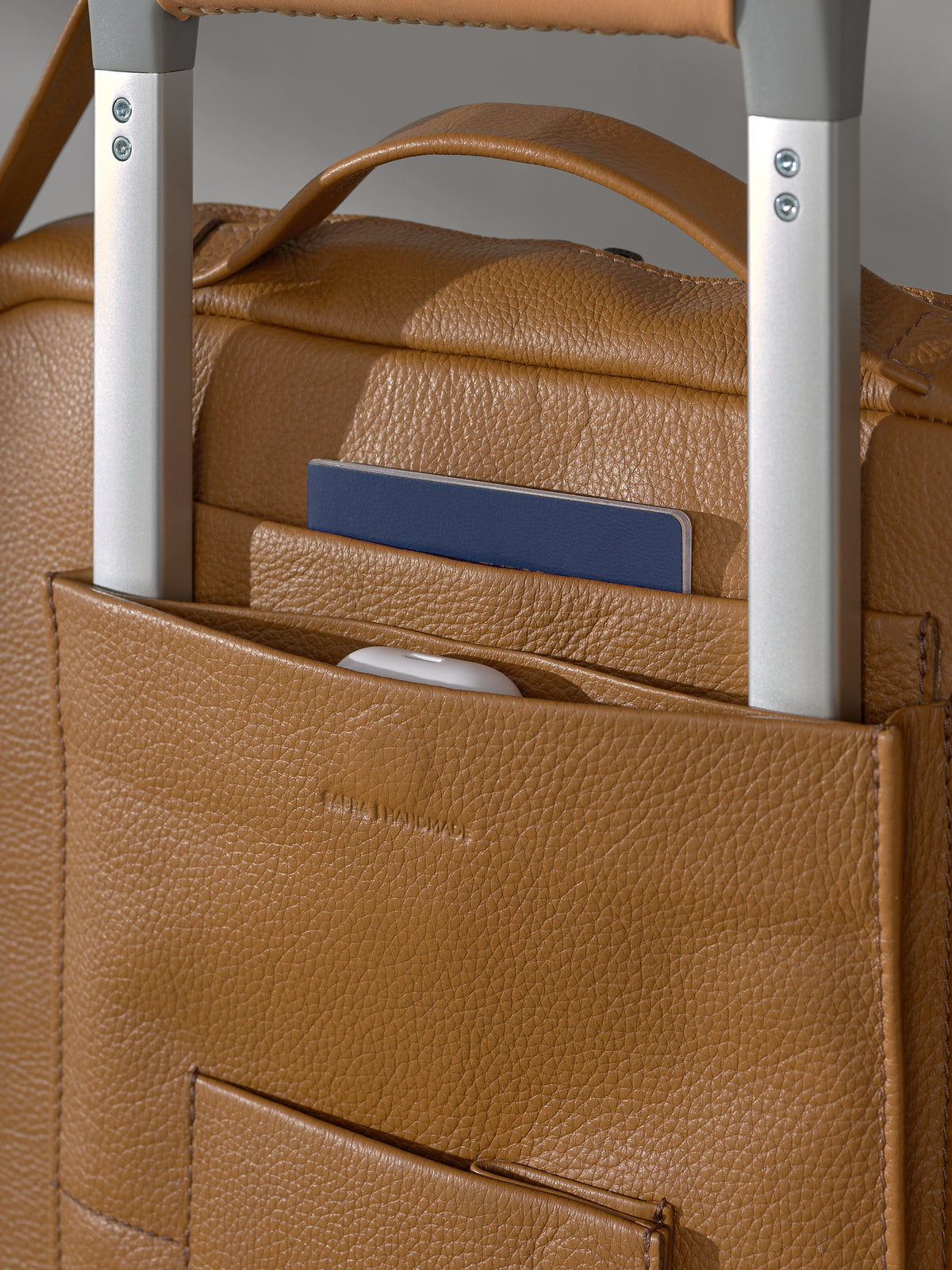 Close-up of the trolley sleeve on the Capra Leather tan travel bag, showcasing dedicated quick-access pockets holding a passport and wireless earbud case.