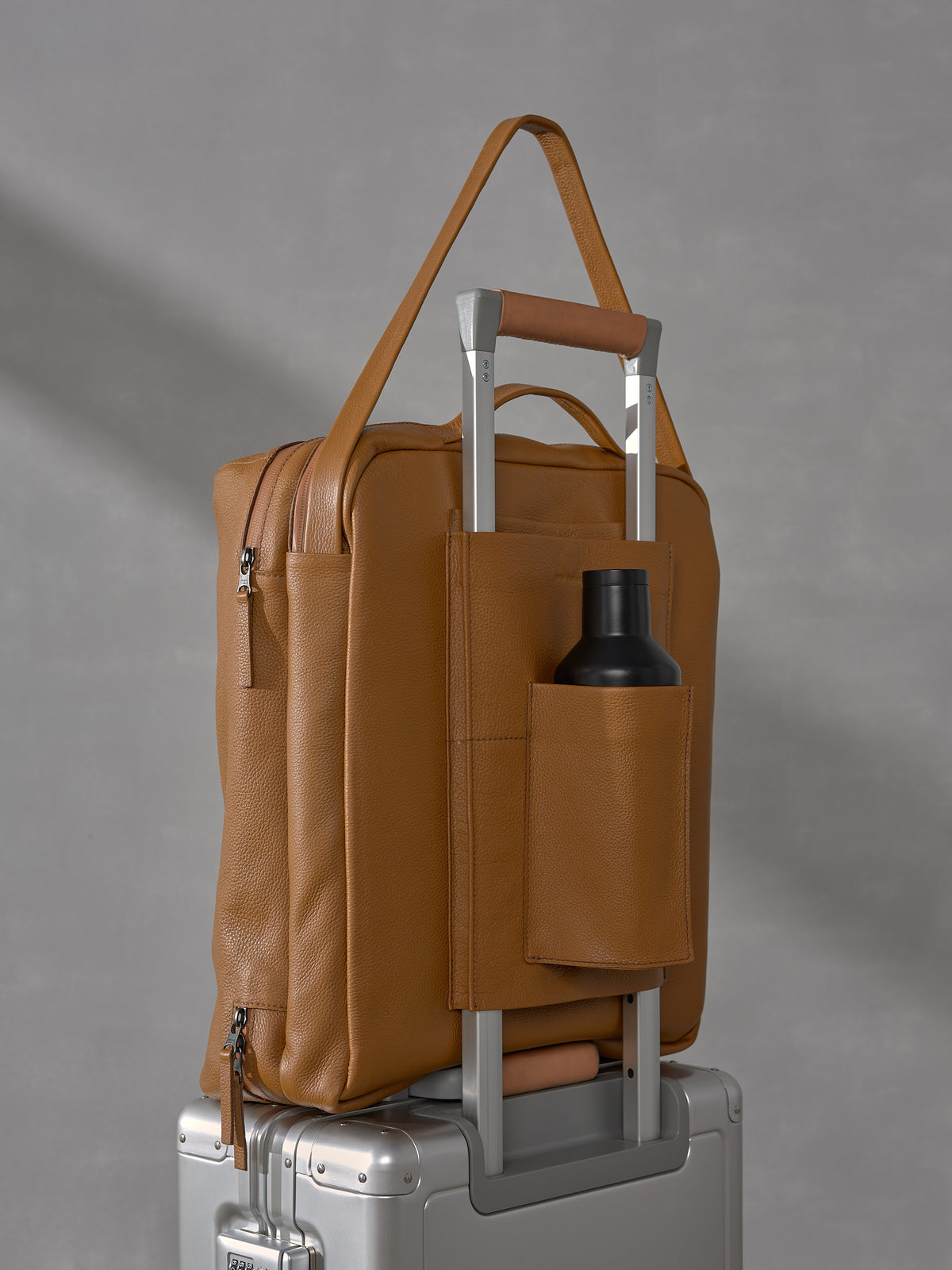 Back view of the Capra Leather tan travel bag showing the integrated trolley sleeve secured over a suitcase handle with a built-in water bottle pocket holding a black flask.
