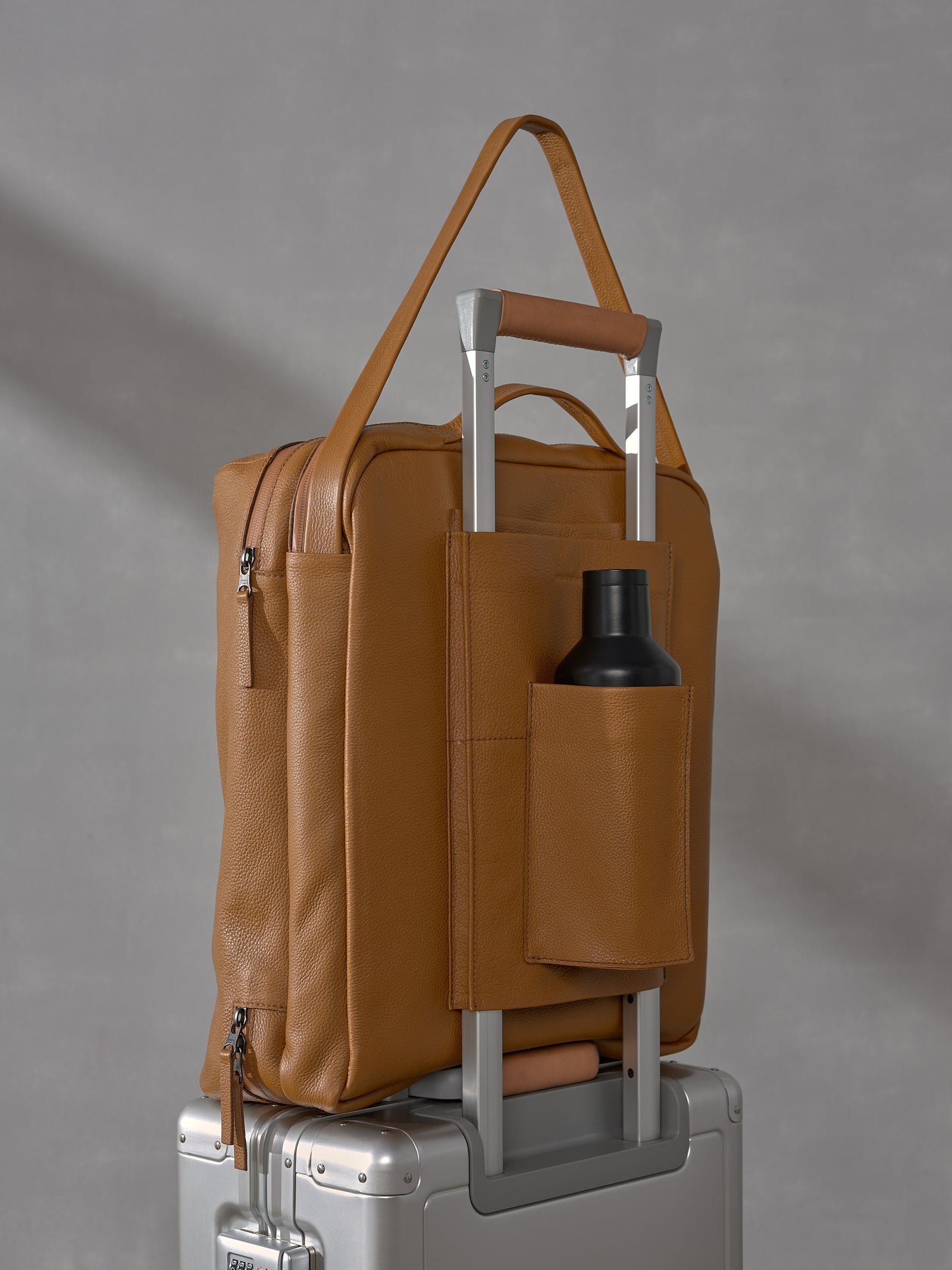 Tan full-grain leather carry-on bag from the Basalt System sitting on top of a sleek silver aluminum rolling suitcase against a gray background.