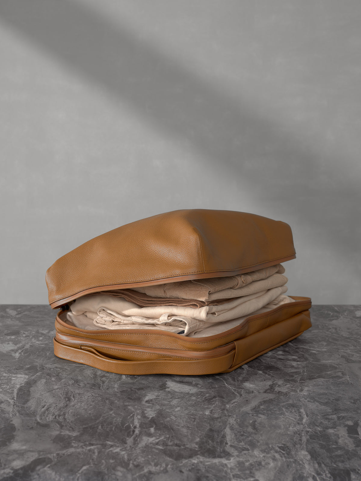 Open tan leather travel organizer pouch from Capra Leather packed with folded cream-colored clothing on a dark marble surface.