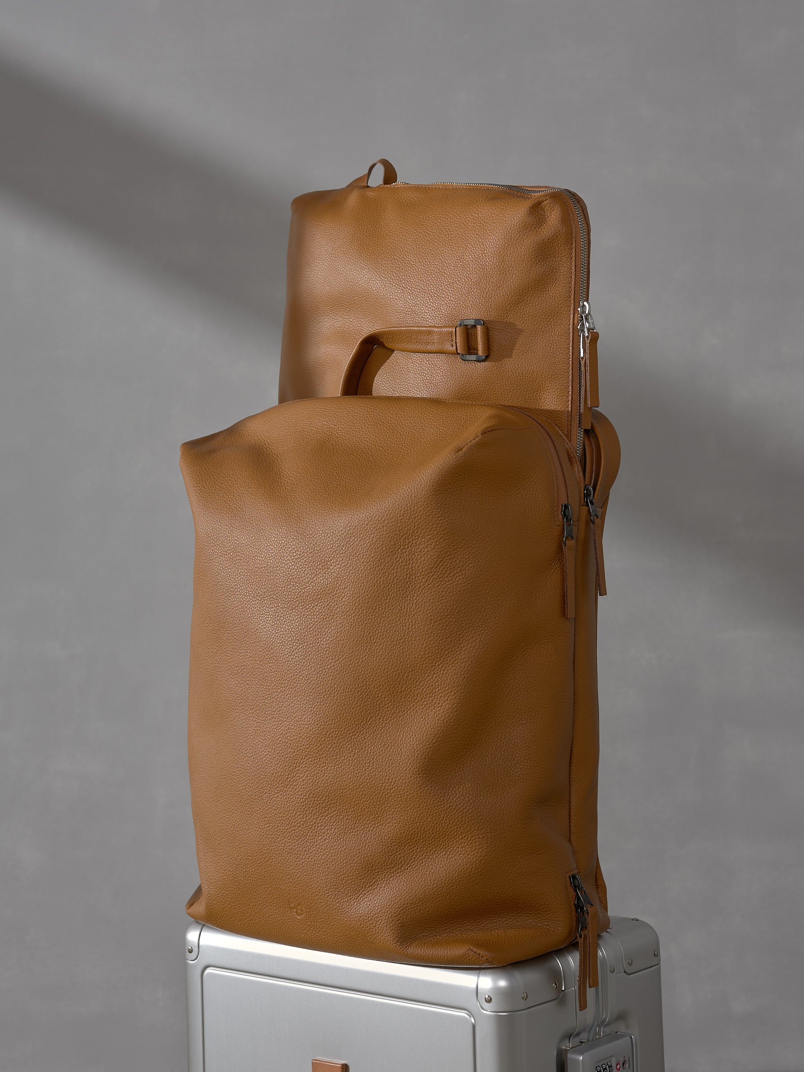 Modular tan leather travel bags from the Capra Leather Basalt system stacked neatly on top of a silver carry-on suitcase.