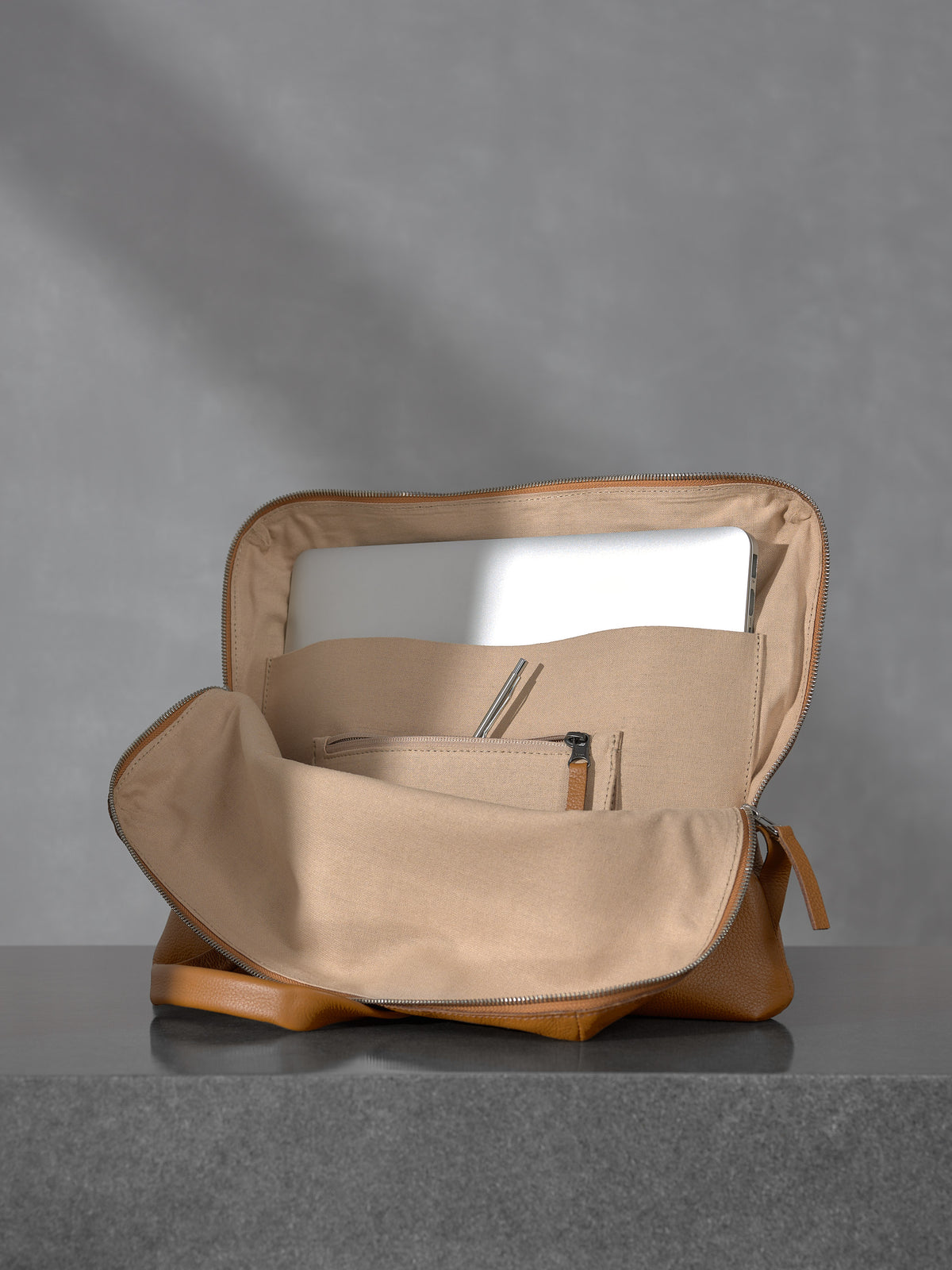 Internal view of the open Capra Leather tan briefcase showing a dedicated laptop compartment holding a silver MacBook, canvas lining, and zippered interior pockets.