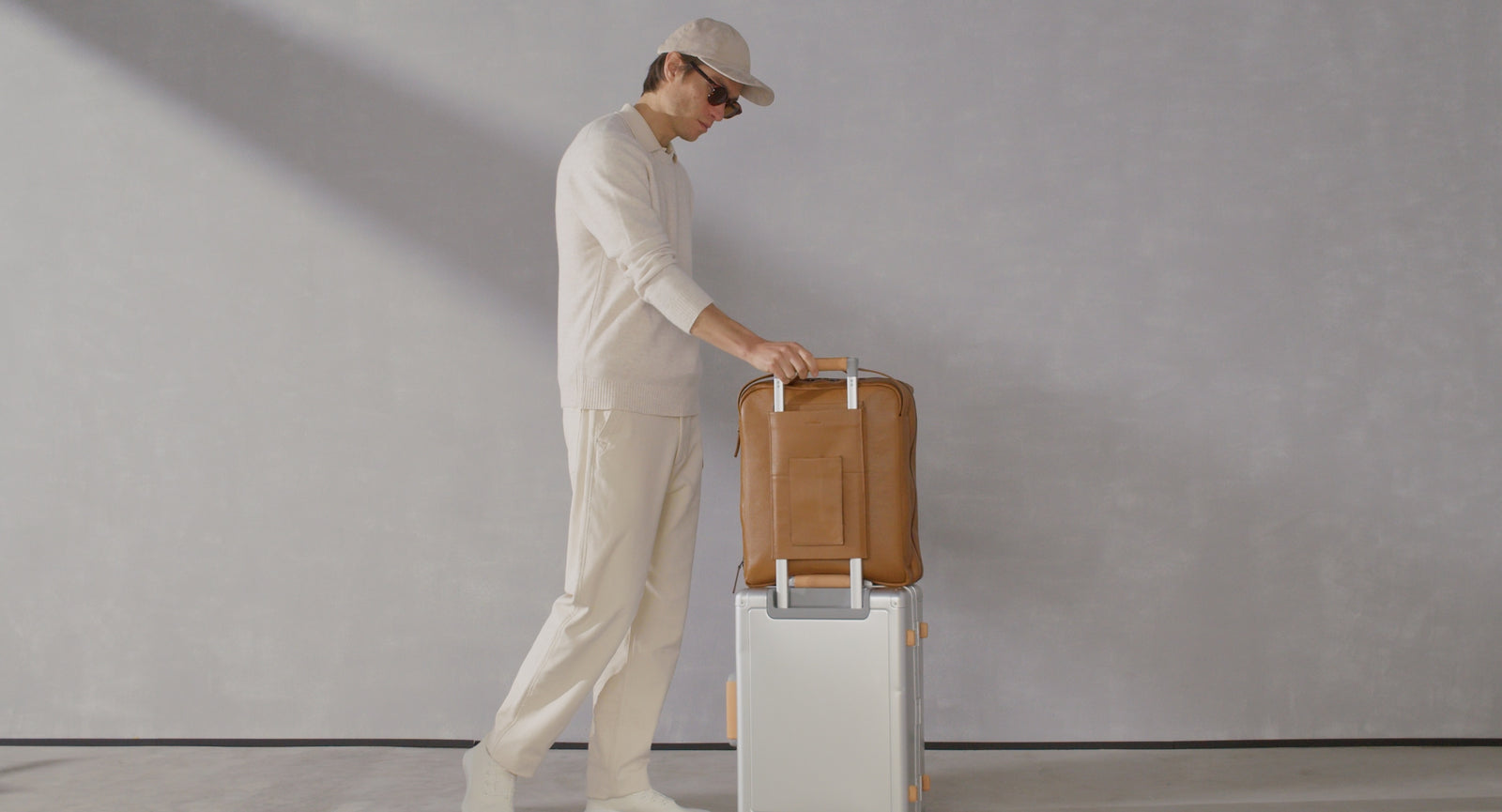 Man in off-white attire walking with a silver rolling suitcase topped with a tan full-grain leather travel bag.