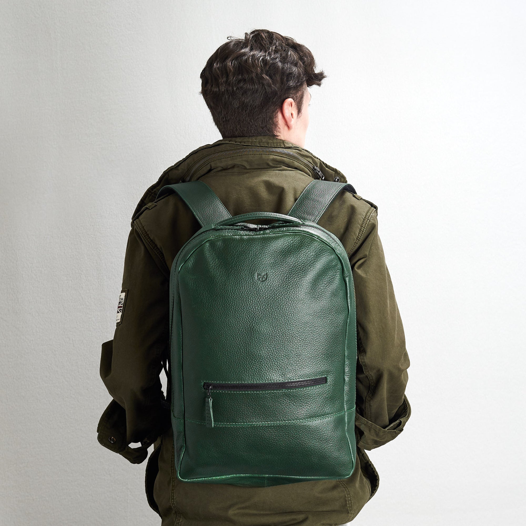A view of a young man's back as he wears the green, pebble-grain leather backpack, showcasing the shoulder straps and the full back panel of the bag.