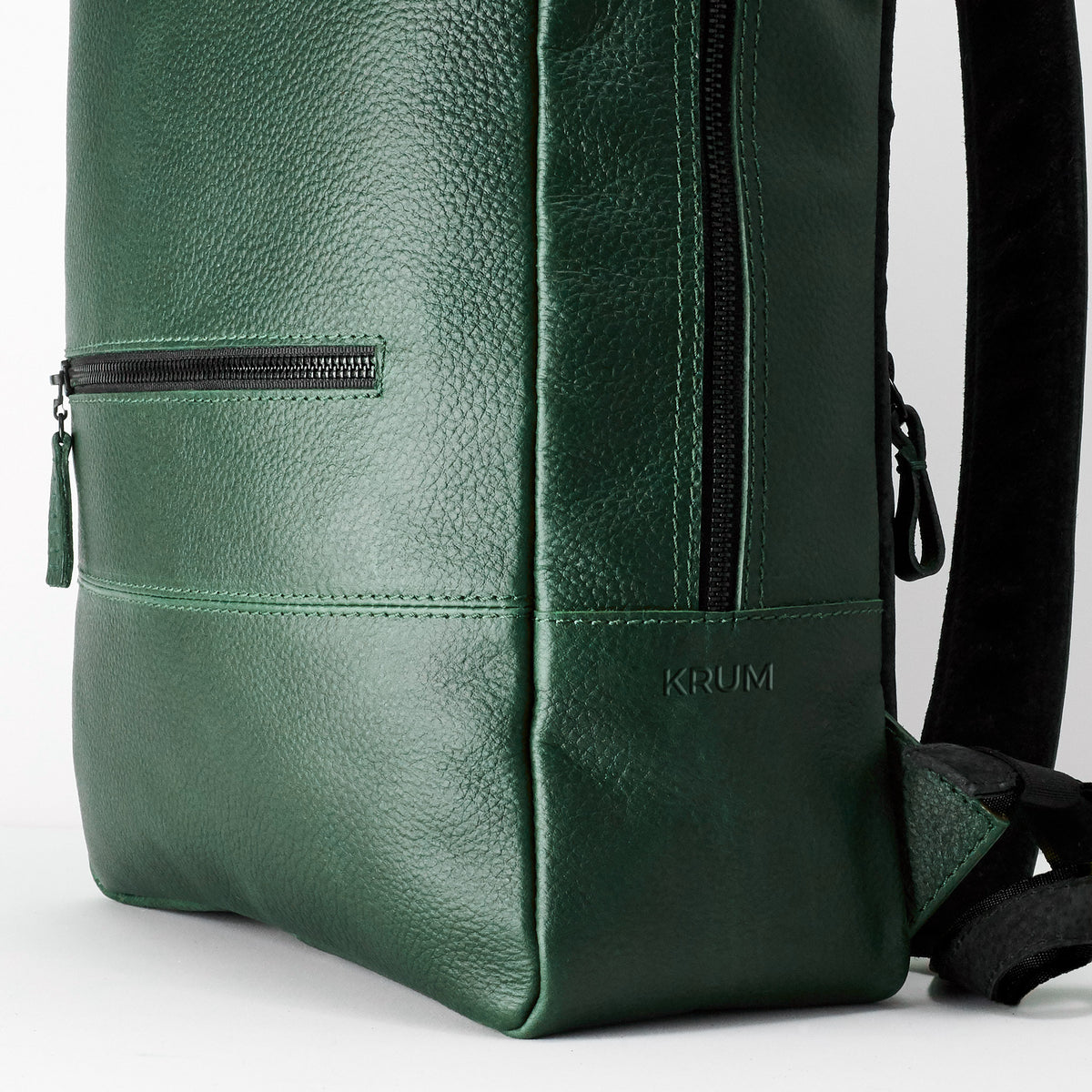 A close-up view of the bottom corner of the green, pebble-grain leather backpack, showing a horizontal zippered pocket, stitching details, and the embossed word "KRUM" near the base.