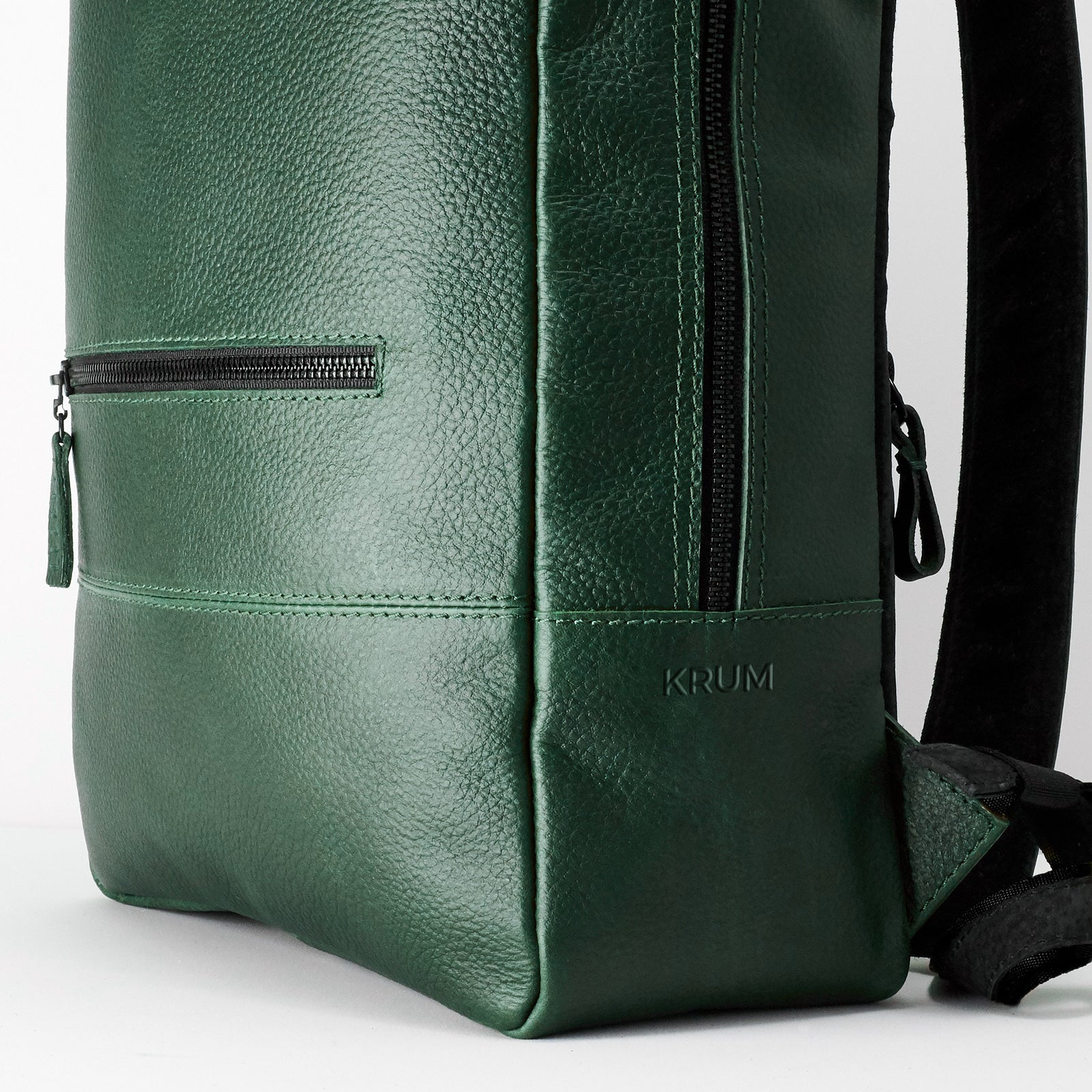 A close-up view of the bottom corner of the green, pebble-grain leather backpack, showing a horizontal zippered pocket, stitching details, and the embossed word "KRUM" near the base.