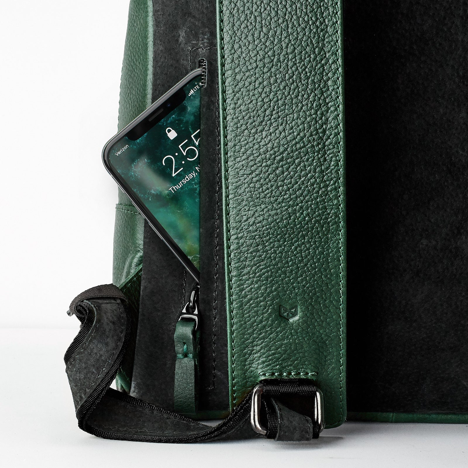 A close-up shot of a narrow, vertically zippered pocket on the side of the green leather backpack, with a smartphone partially tucked inside.