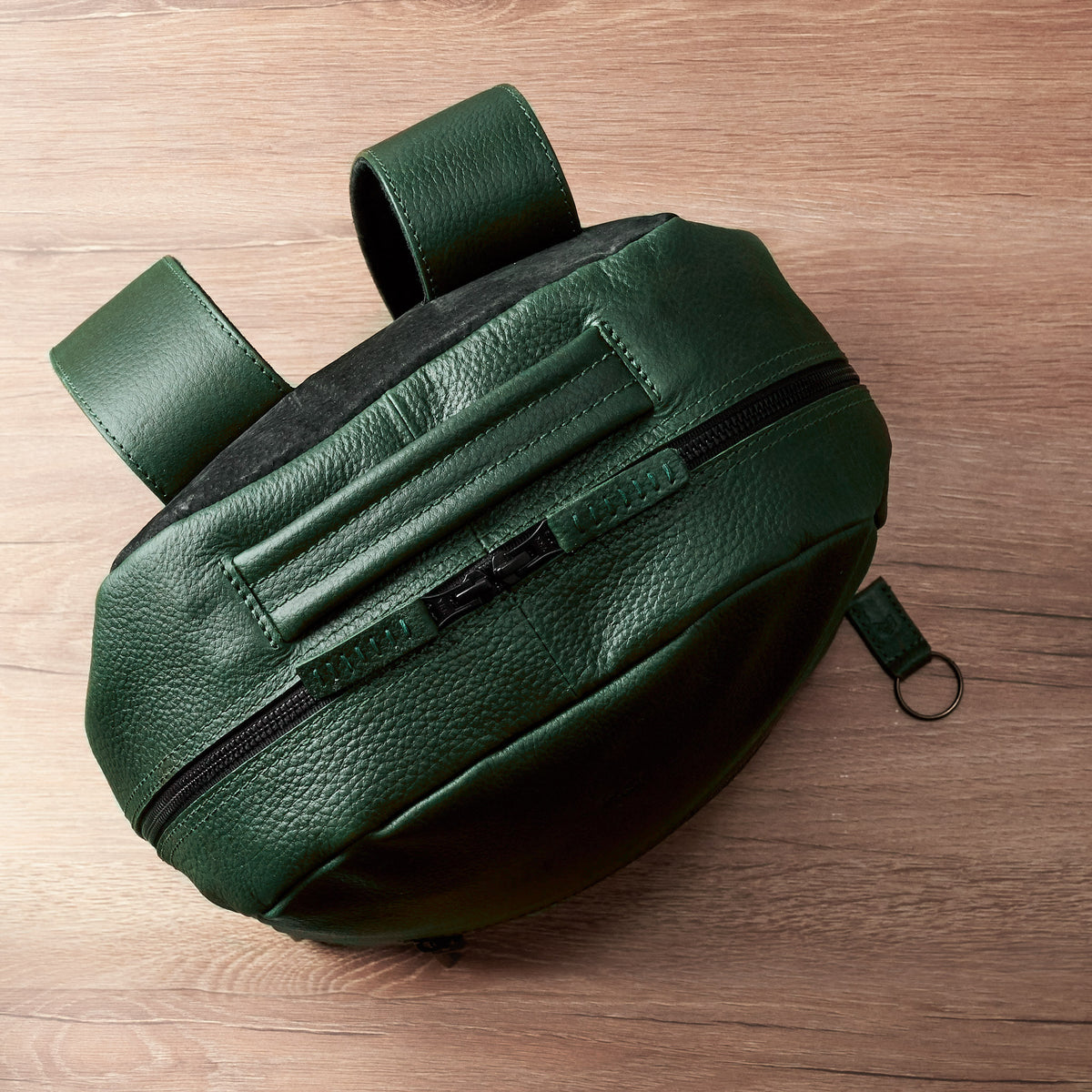An overhead shot of the rounded top of the green, pebble-grain leather backpack lying on a wooden surface, showing the zipper running along the top edge and the attached leather shoulder straps.