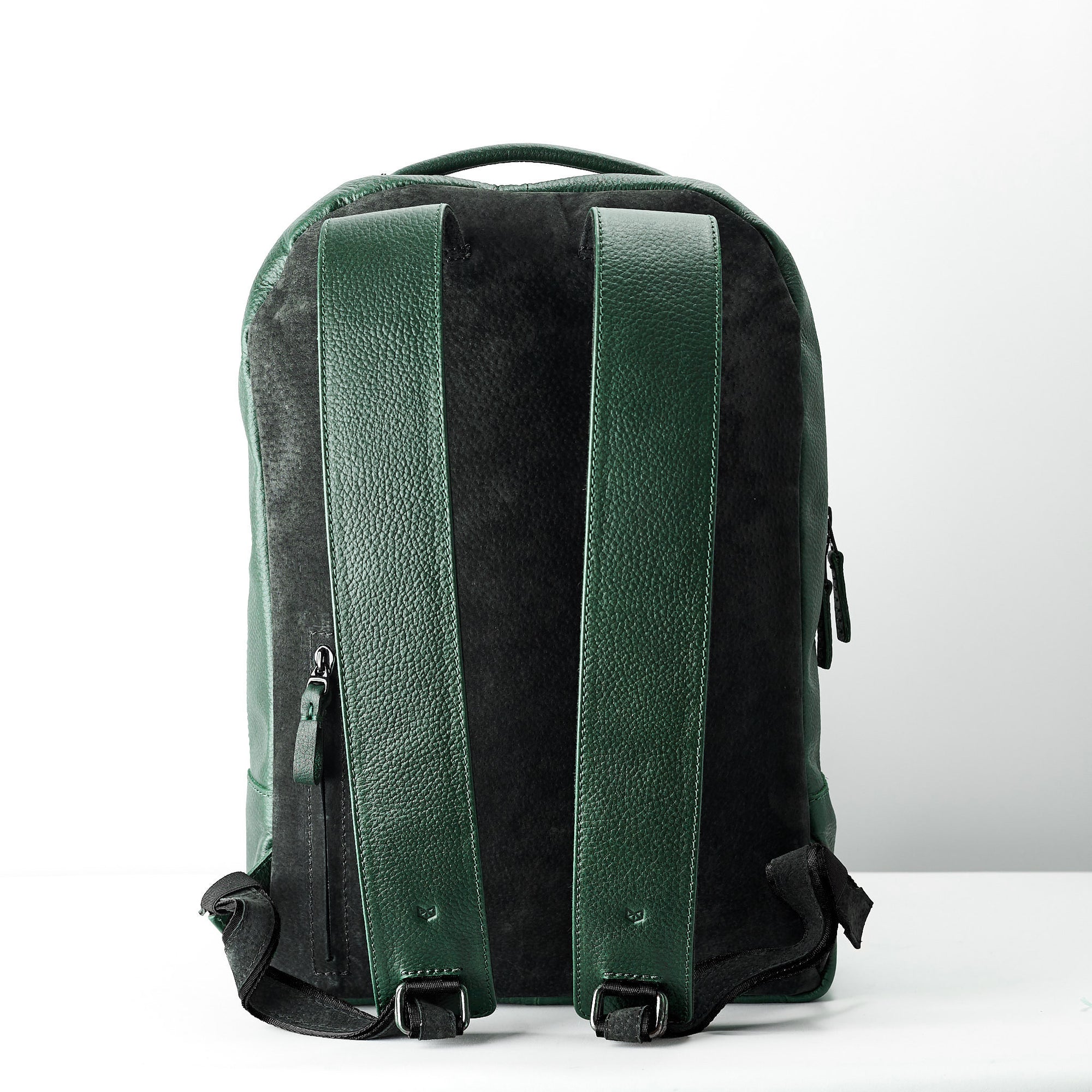 The back view of the green leather backpack, showing two wide, adjustable shoulder straps made of green leather and the contrasting dark suede/fabric back panel. A small zippered pocket is visible on the left side.