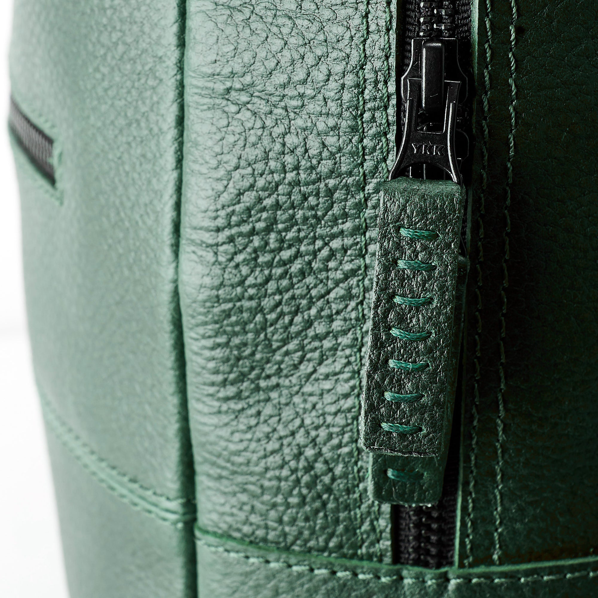 An extreme close-up detail of the black YKK zipper and the green leather zipper pull tab with visible dark green stitching on the pebble-grain leather backpack.
