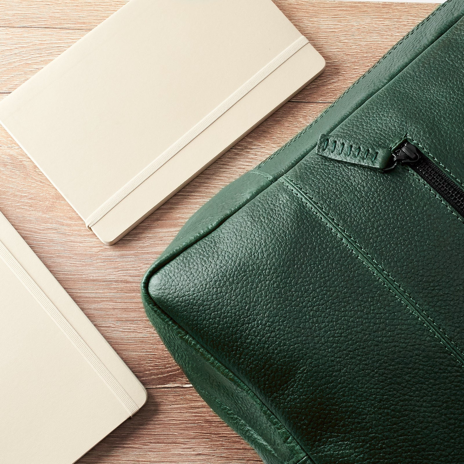 A close-up flat lay shot of the corner of the green leather backpack next to two cream-colored notebooks on a wooden surface.