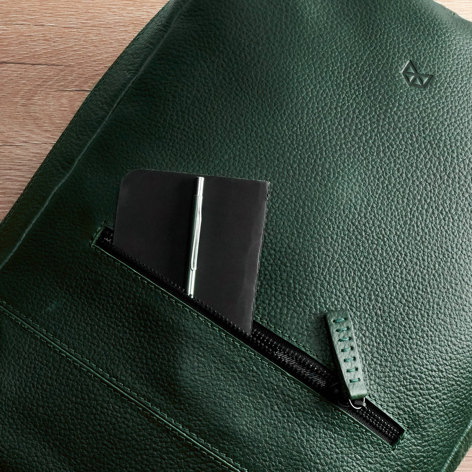 A very close-up detail of the front zipper pocket on the green leather backpack, which holds a small black notebook and a pen.