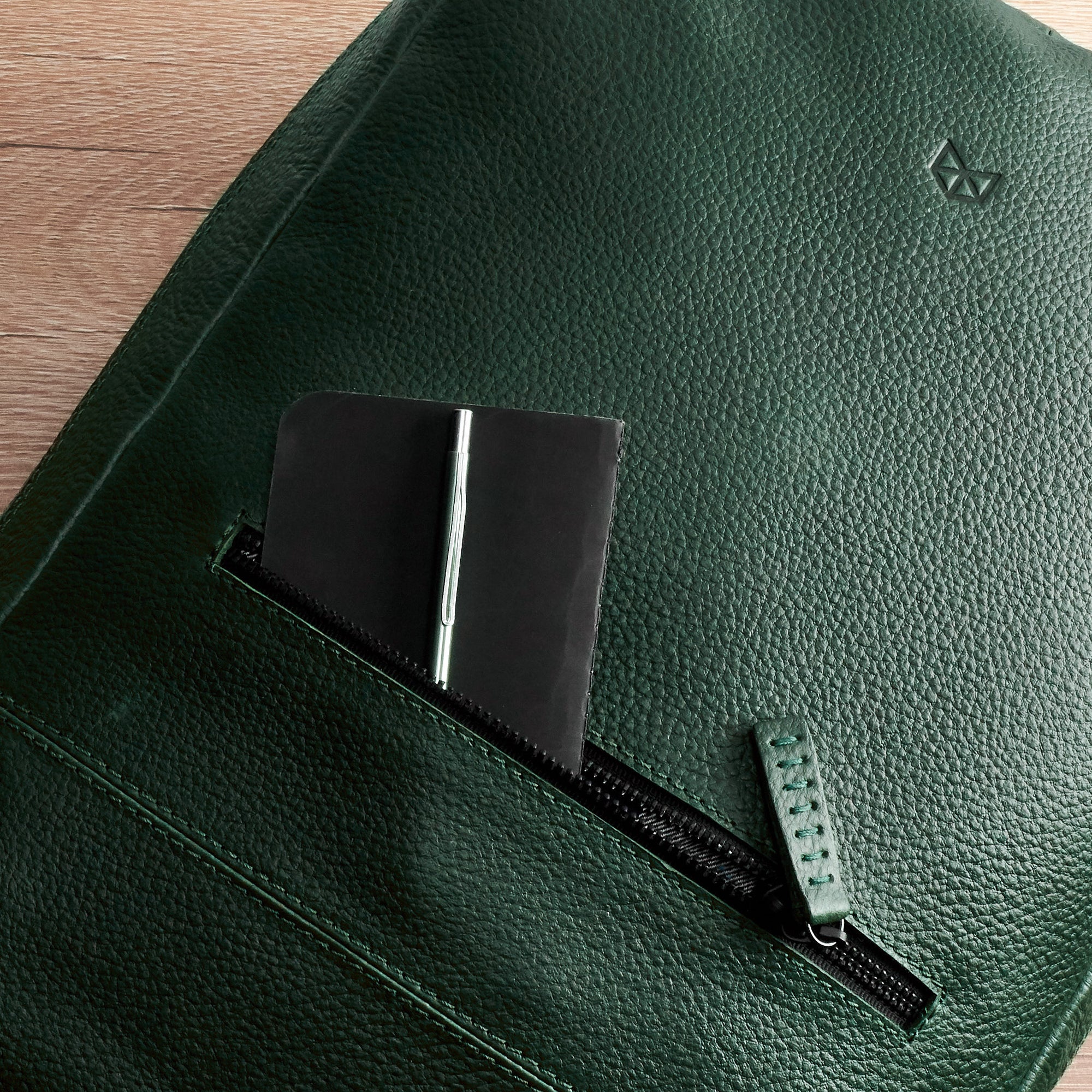 A very close-up detail of the front zipper pocket on the green leather backpack, which holds a small black notebook and a pen.