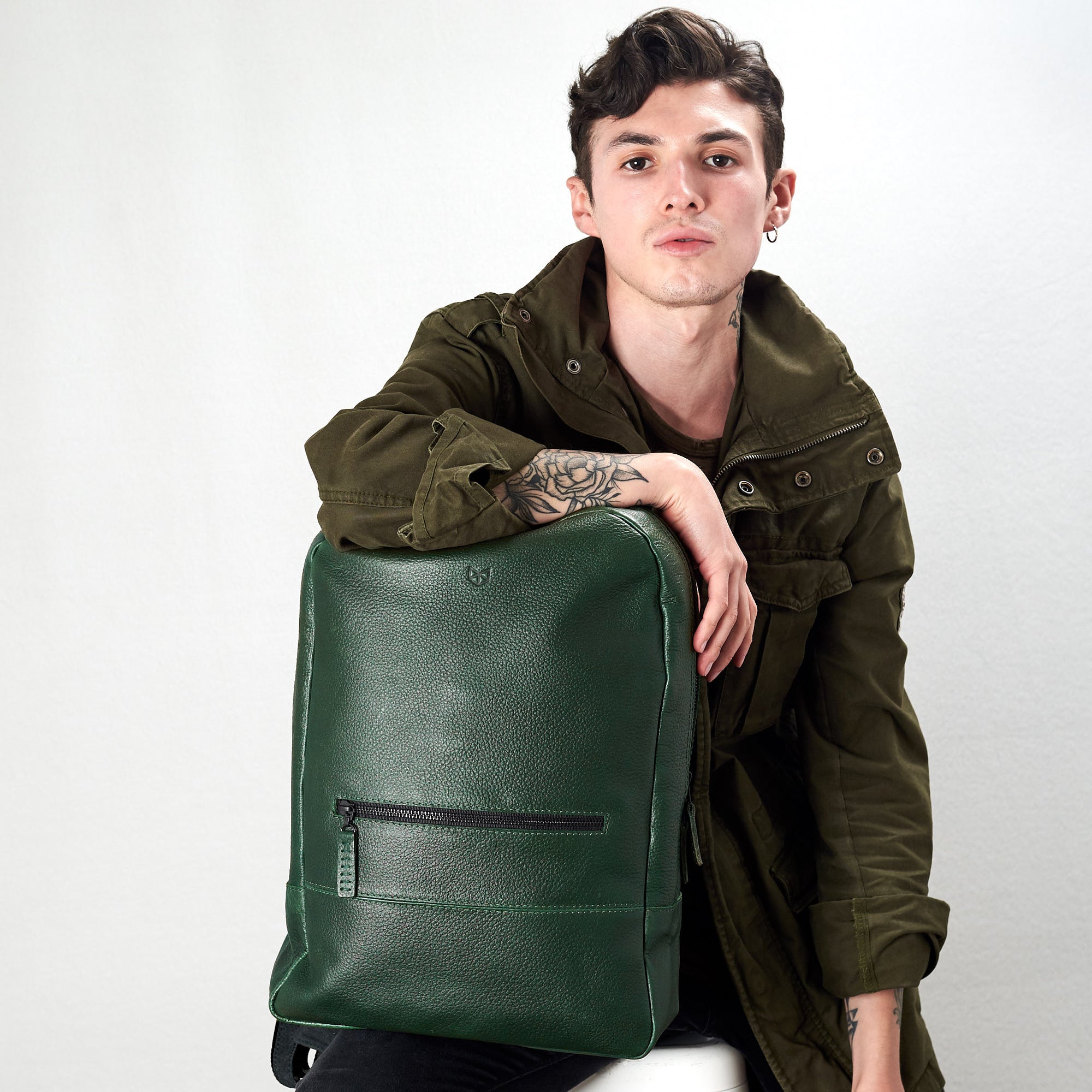 A young man with tattoos, wearing an olive green jacket, sitting and leaning on the green, pebble-grain leather backpack, which is standing upright.