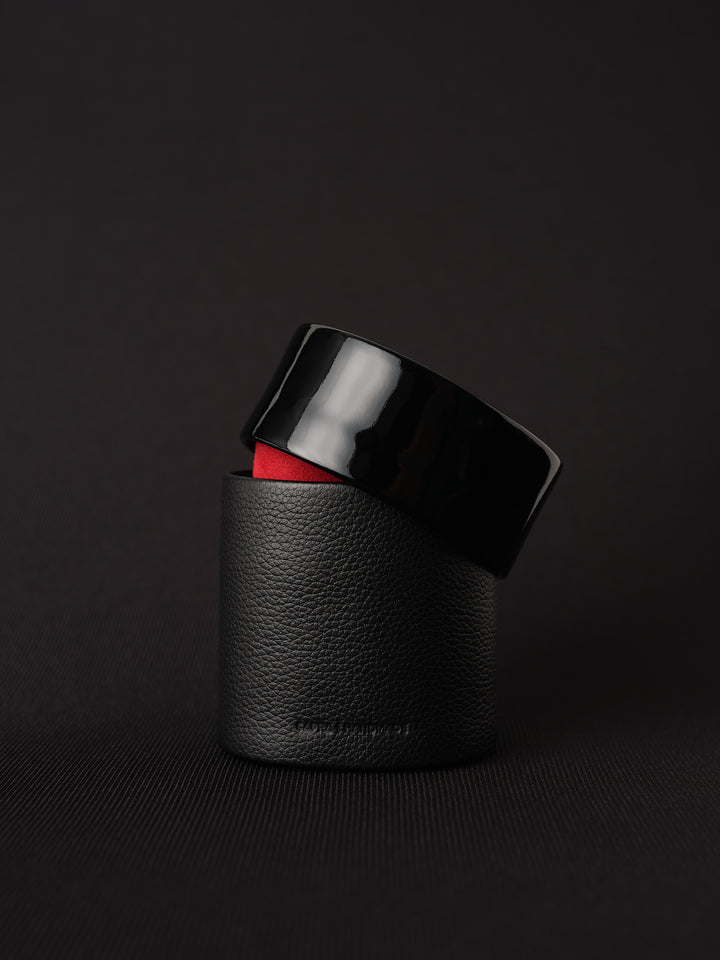 A single short, cylindrical black watch case on a black textured surface. The case has a pebbled leather base and a glossy black lid, which is slightly ajar, revealing the red inner lining.