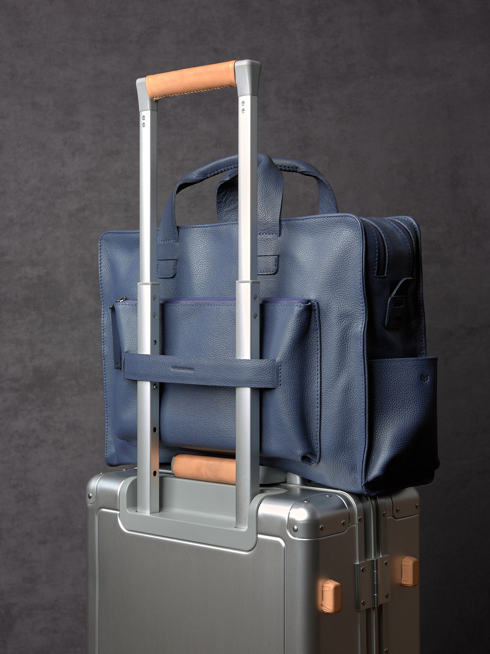 Capra Leather navy blue briefcase attached to a silver rolling suitcase handle using its built-in luggage strap.