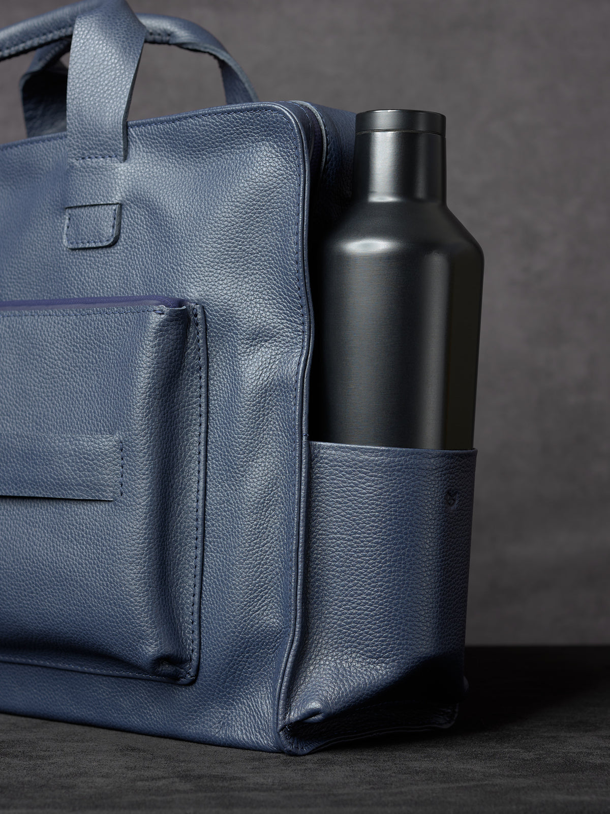 Side view of Capra Leather navy blue leather bag showing a black water bottle tucked into the side pocket.