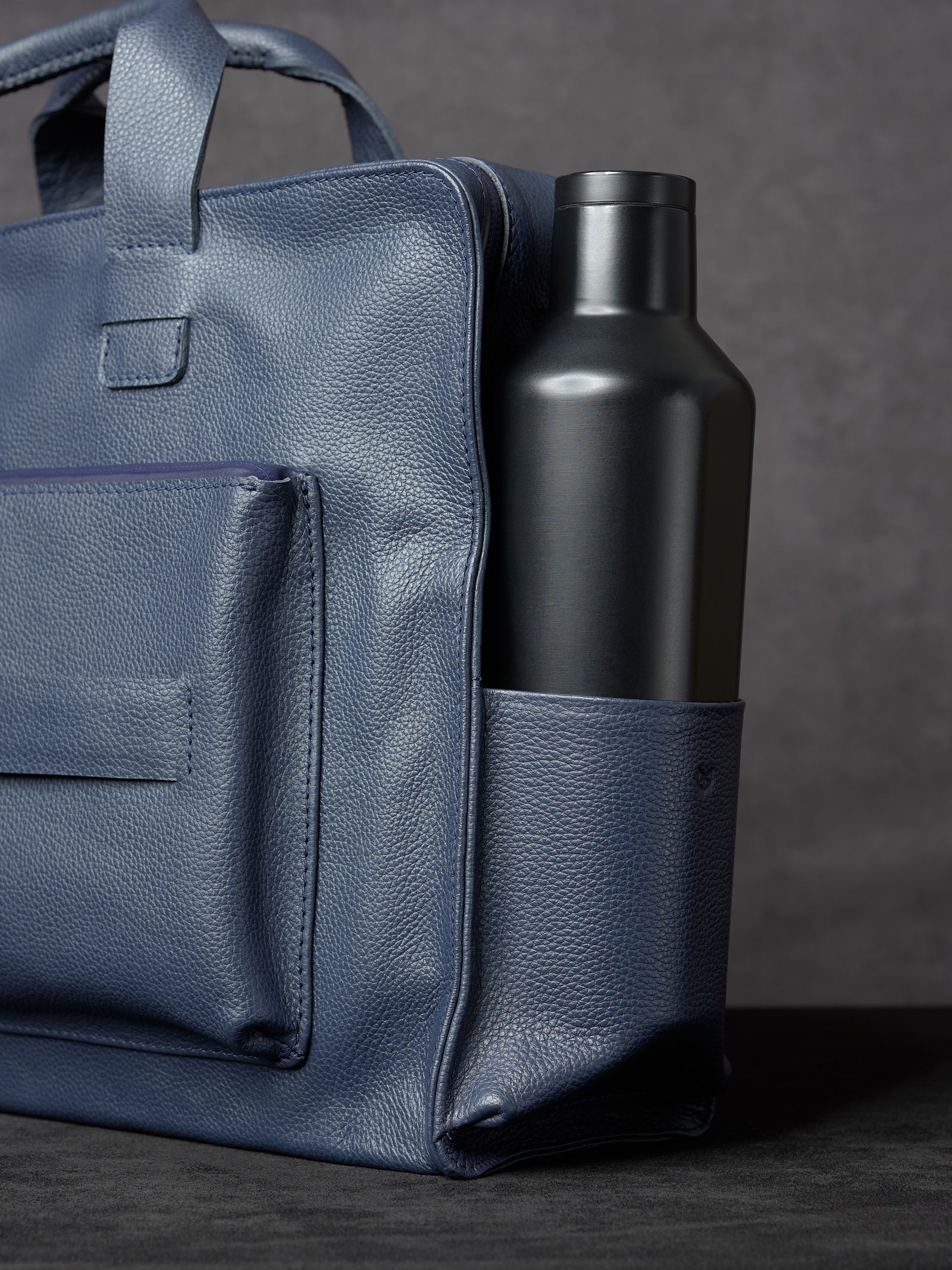 Side view of Capra Leather navy blue leather bag showing a black water bottle tucked into the side pocket.