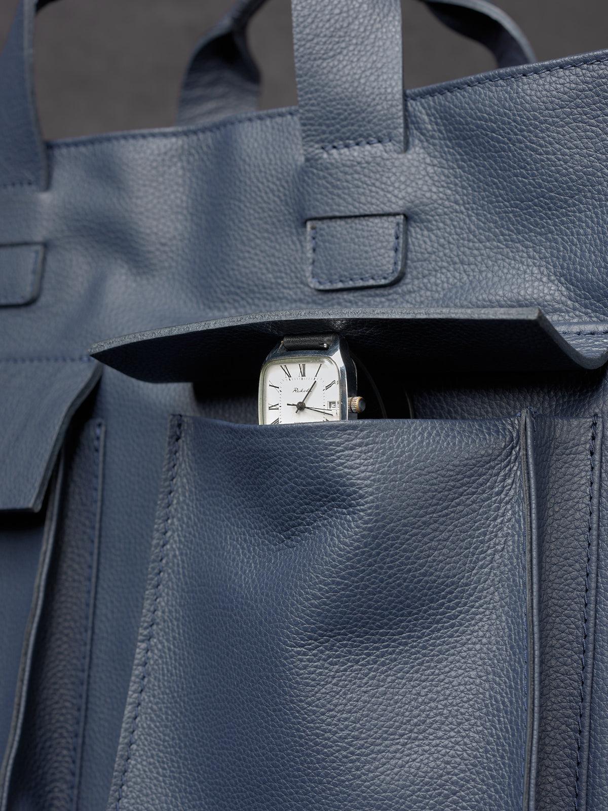 Close-up of a silver wristwatch tucked into the front pocket of a navy blue leather Capra Leather bag.