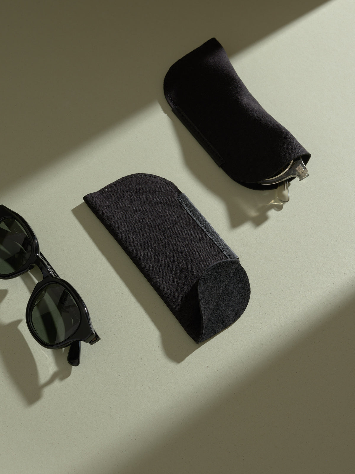 Two soft black pouches for the double-glasses case, one holding clear frames, next to black sunglasses.