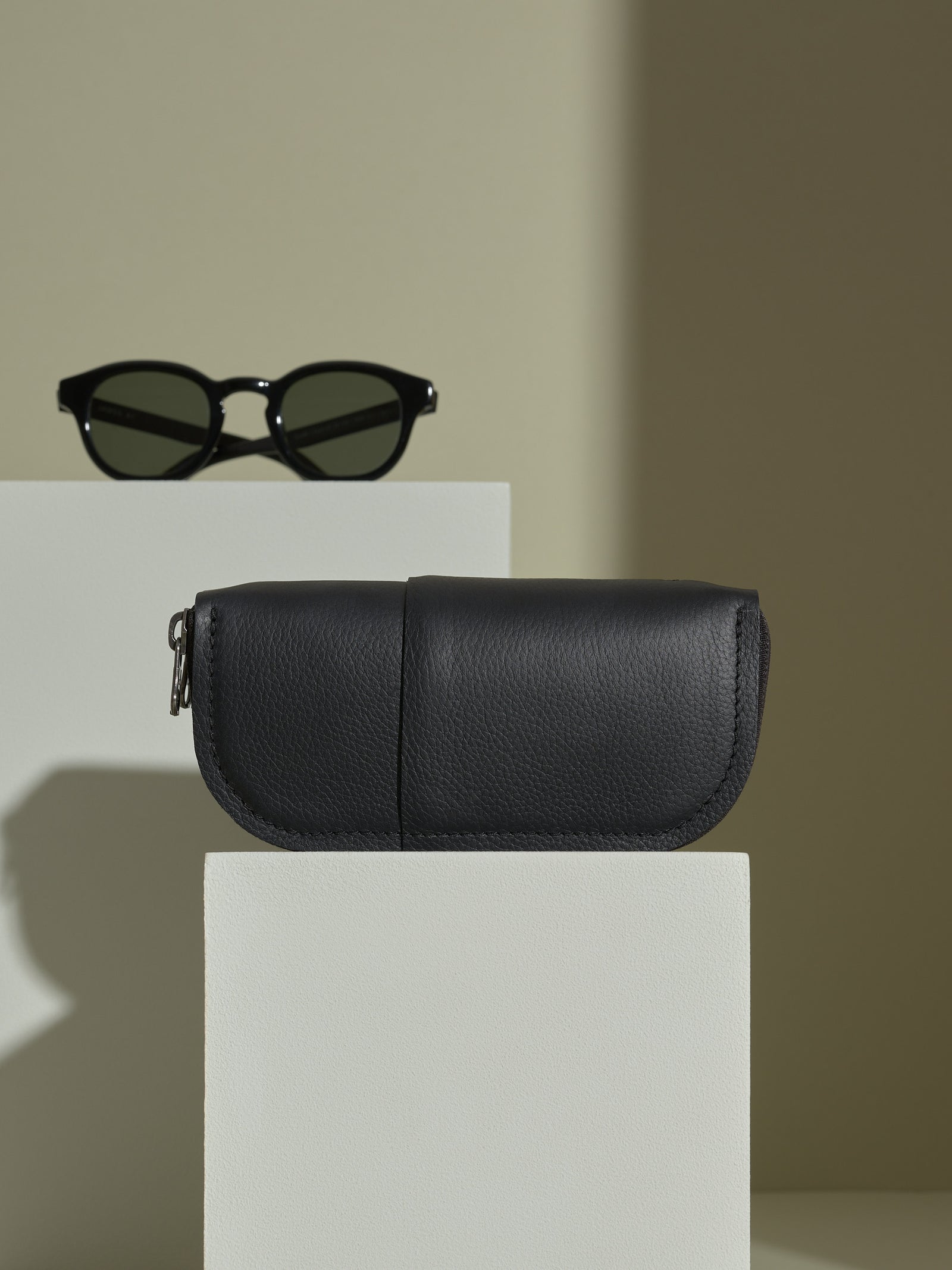 Closed, premium black leather double-glasses case displayed on a white block with black sunglasses on a separate riser.