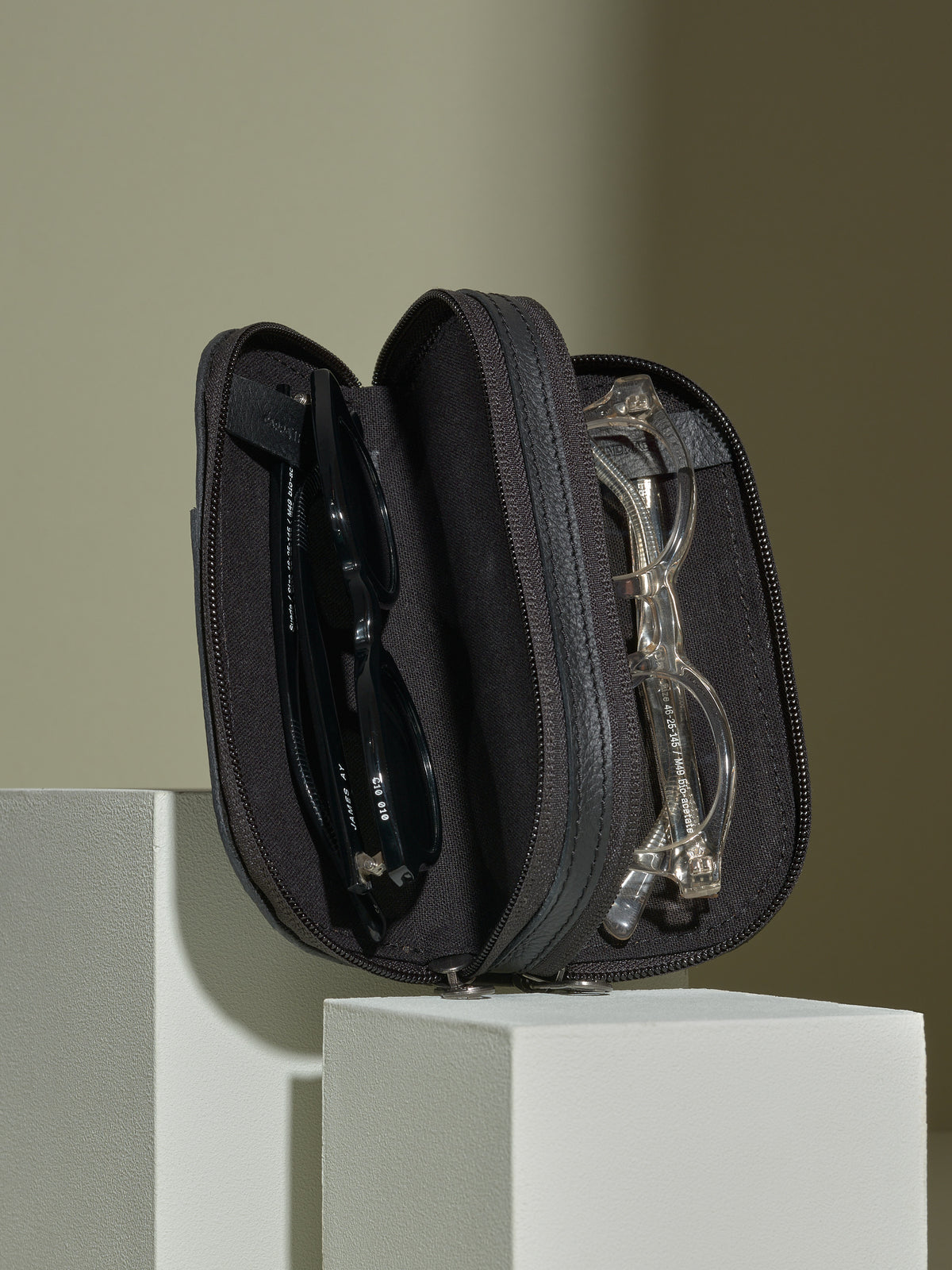Black leather double-glasses case opened on white blocks, showing a pair of sunglasses and reading glasses inside.