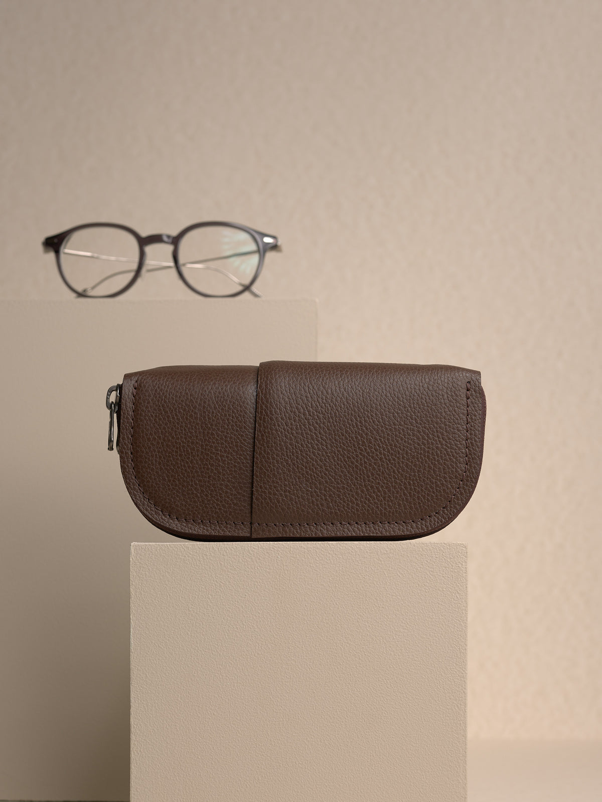 The closed, brown Capra leather double glasses case resting horizontally on two cream-colored platforms, with a pair of round, black-framed glasses displayed in the background.