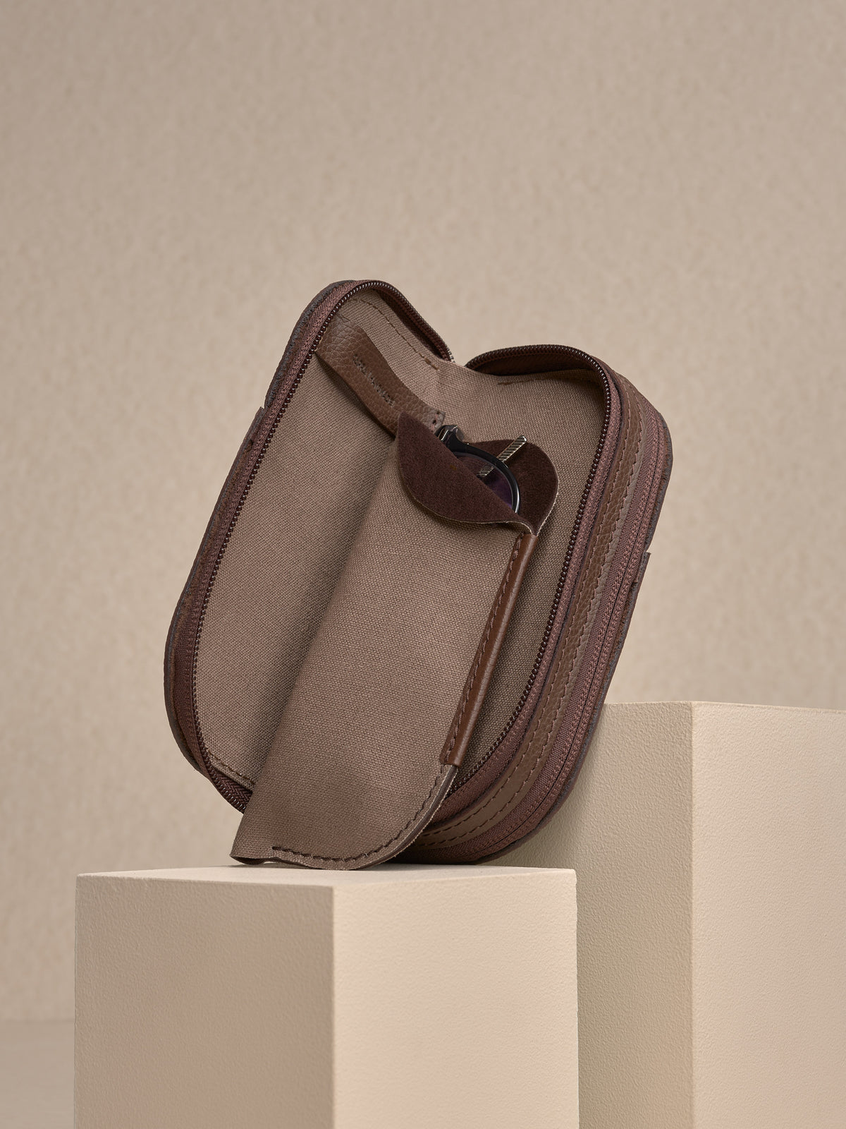 The opened, brown leather zipped glasses case resting on white platforms, with one of the soft brown fabric inner sleeves pulled out and containing a pair of silver-framed glasses.
