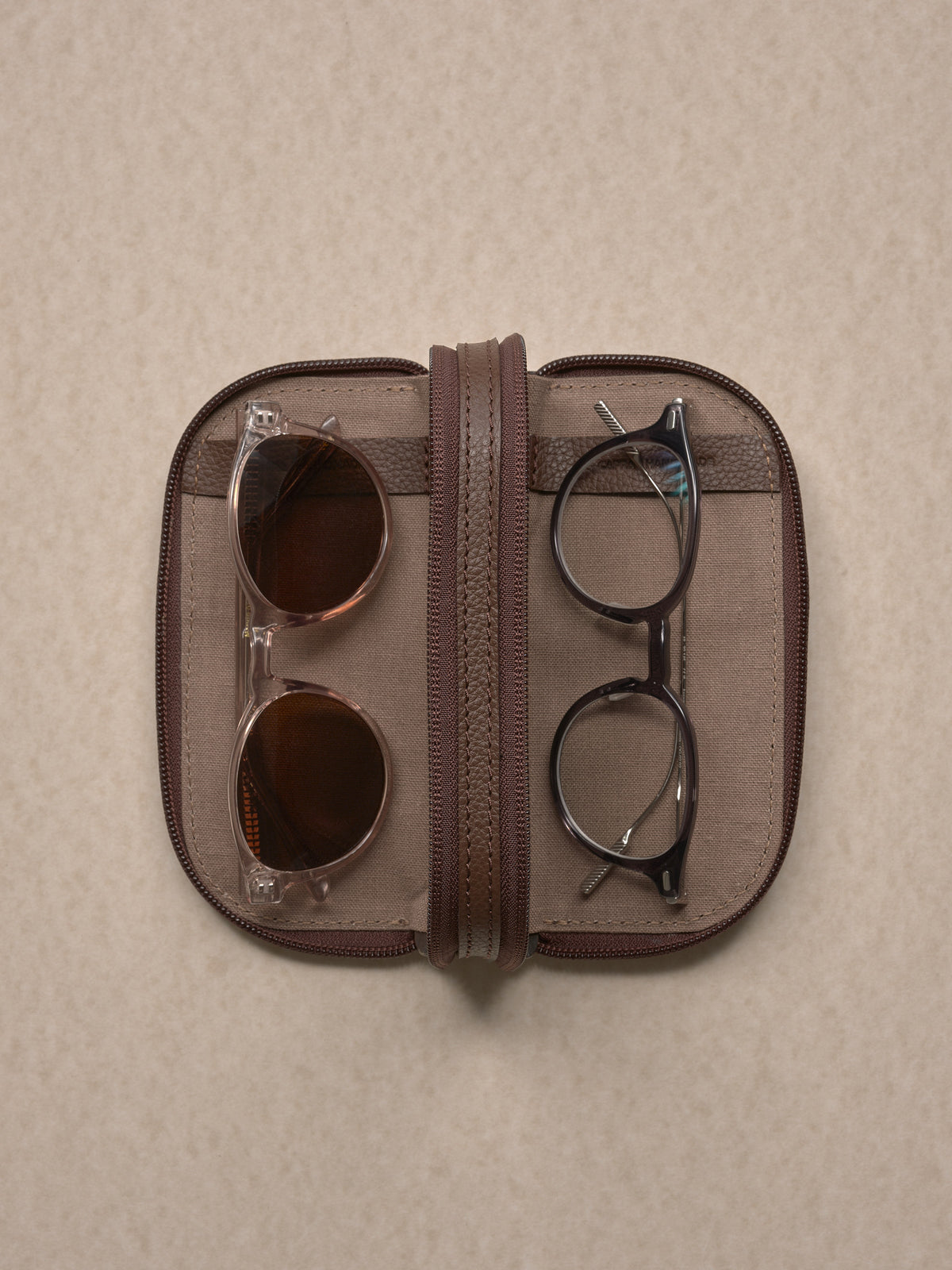 A top-down view of the brown leather glasses case opened, displaying two pairs of glasses secured inside: clear-framed sunglasses on the left and black-framed reading glasses on the right