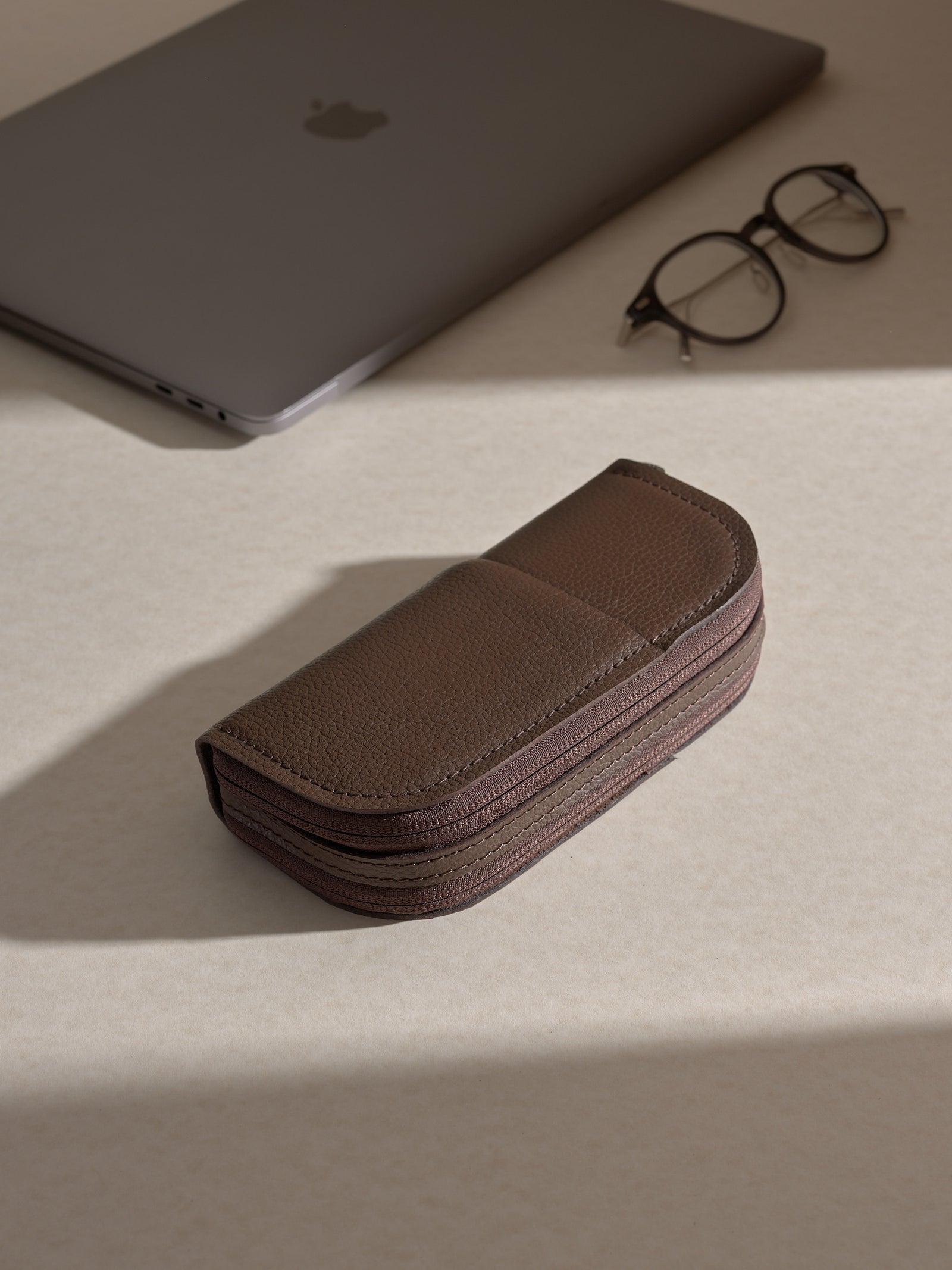 The closed, brown Capra leather double glasses case resting horizontally on two cream-colored platforms, with a pair of round, black-framed glasses displayed in the background.