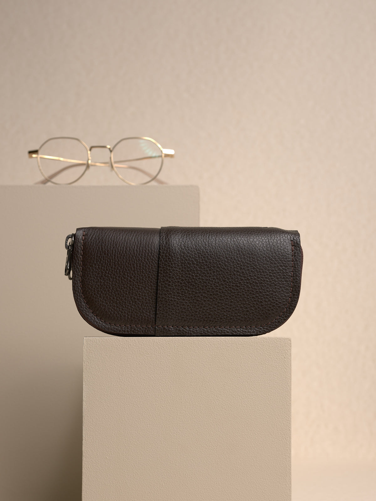The closed, dark brown Capra leather double glasses case resting horizontally on a small light-colored platform, with a pair of gold polygonal-framed glasses in the blurry background.