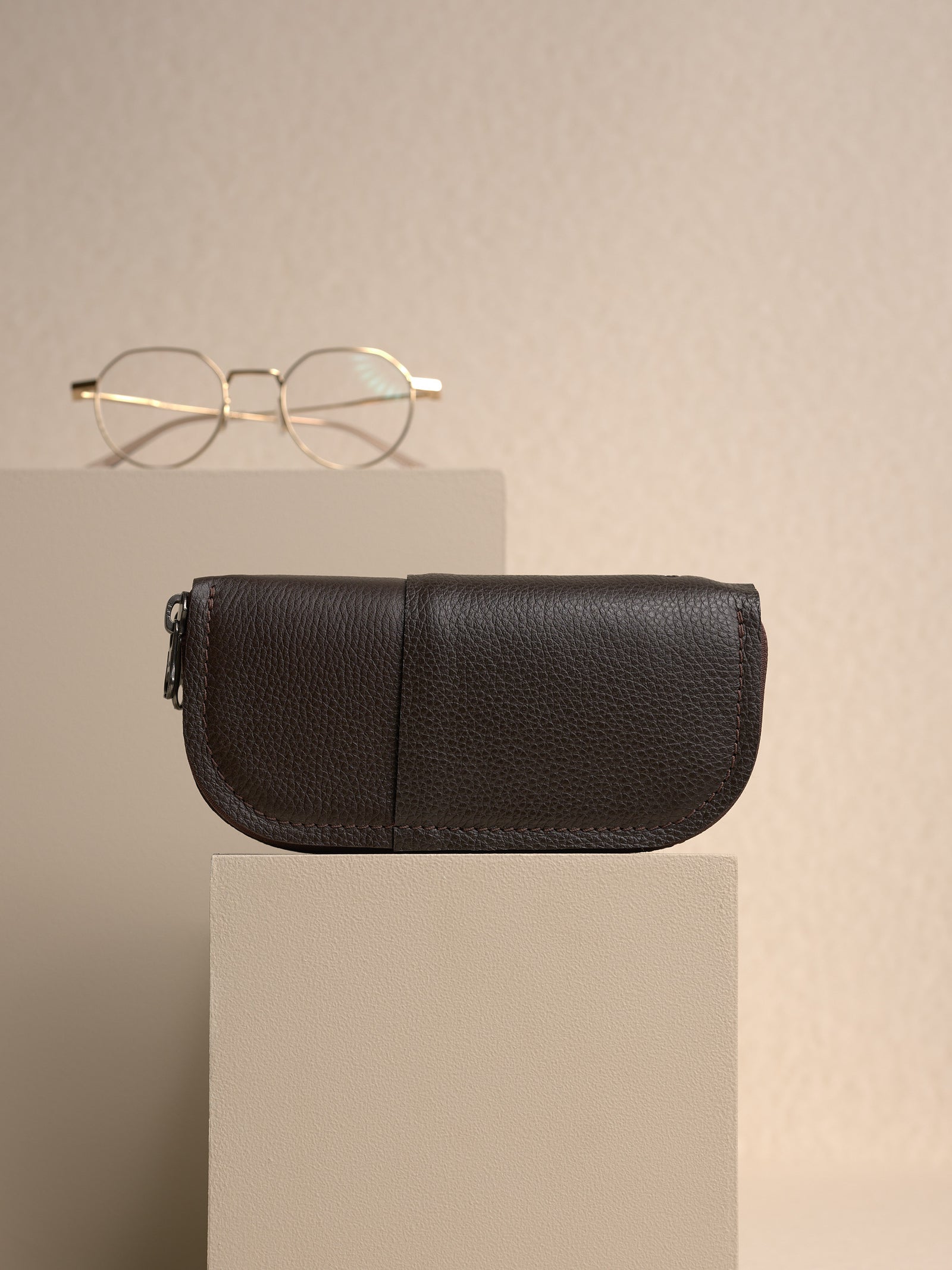 The closed, dark brown Capra leather double glasses case resting horizontally on a small light-colored platform, with a pair of gold polygonal-framed glasses in the blurry background.
