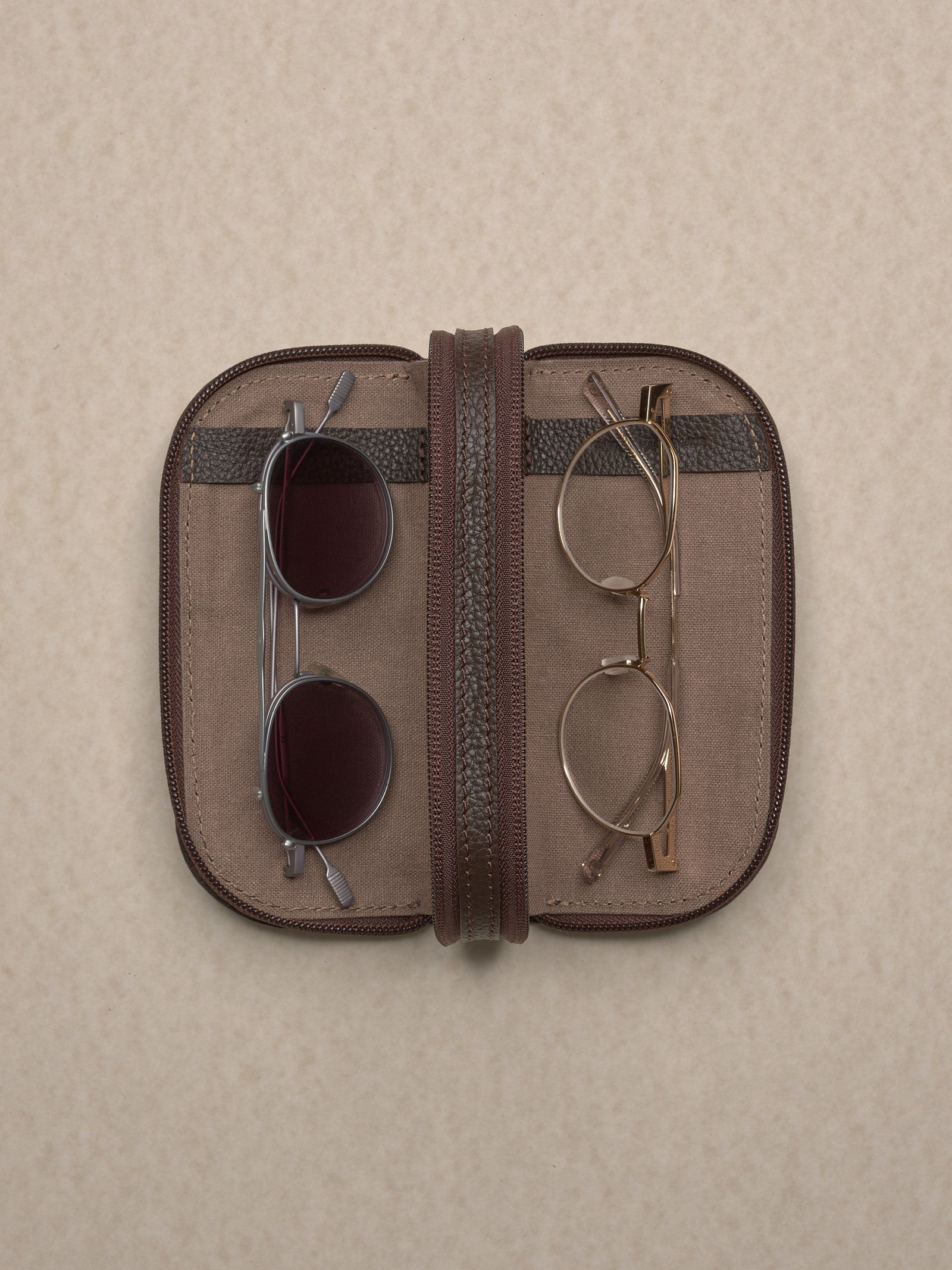 Overhead view of the opened, dark brown leather glasses case, displaying two pairs of glasses (sunglasses on the left, clear-framed on the right) held within the inner compartments.