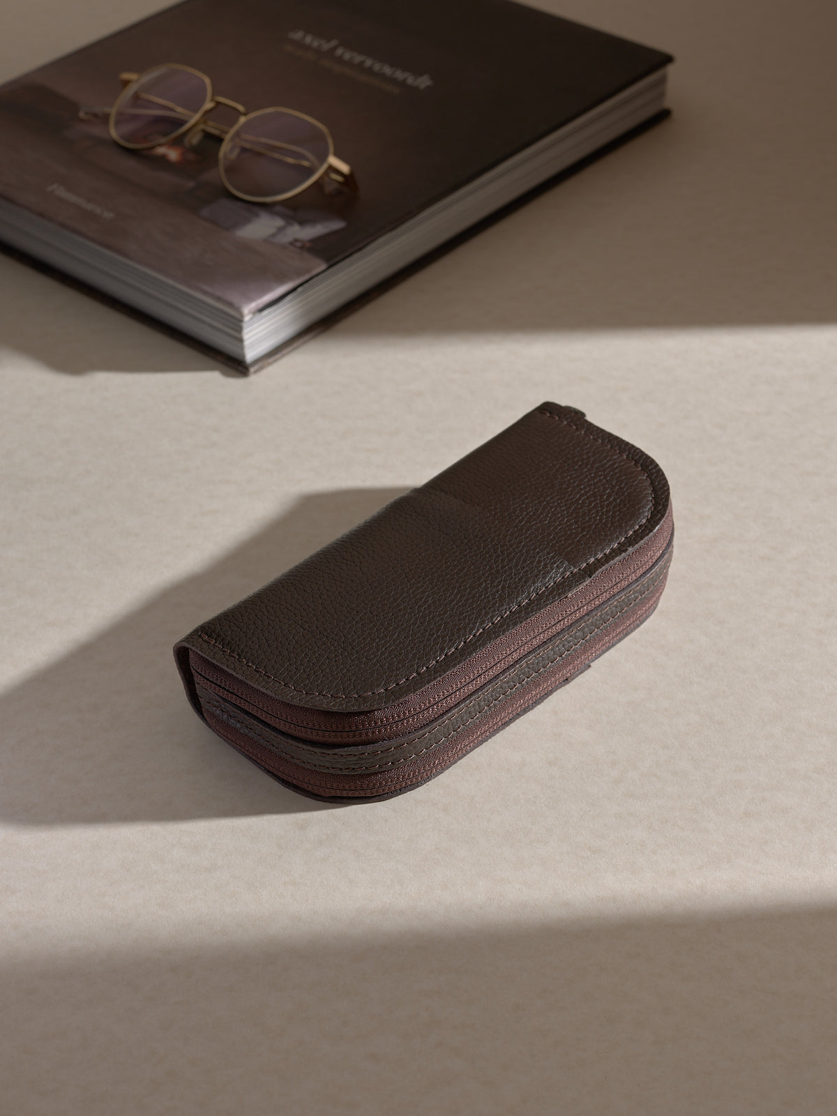 he closed, dark brown Capra leather double glasses case resting on a light-colored desk, next to a gold-framed pair of glasses on top of a book.