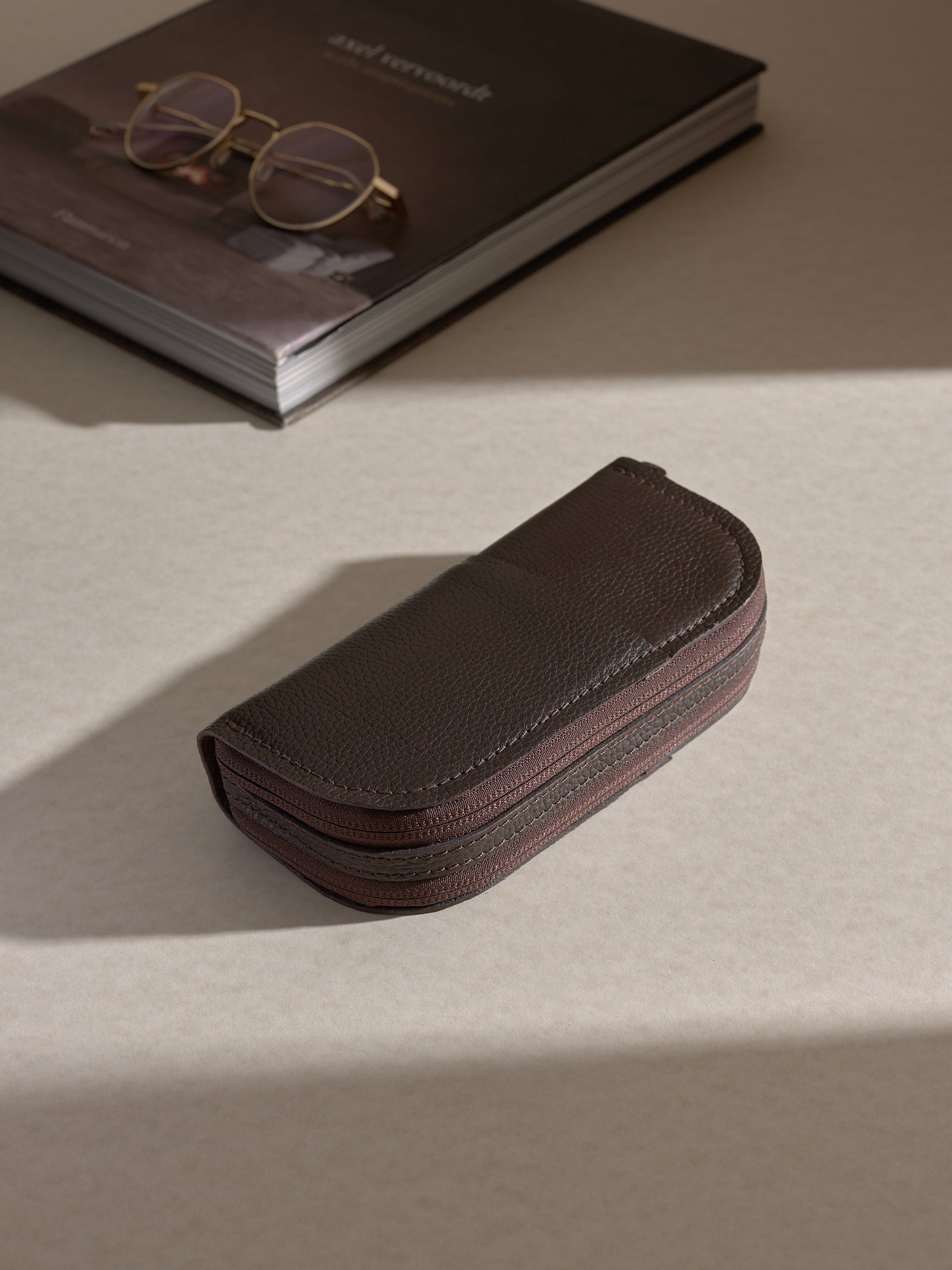 he closed, dark brown Capra leather double glasses case resting on a light-colored desk, next to a gold-framed pair of glasses on top of a book.