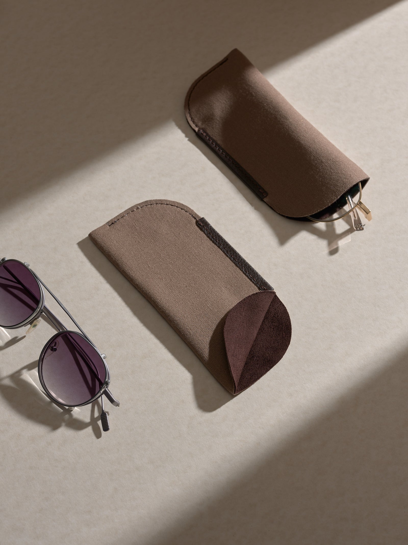 Two separate soft dark brown fabric inner sleeves displayed on a surface, next to a pair of silver-framed round sunglasses.