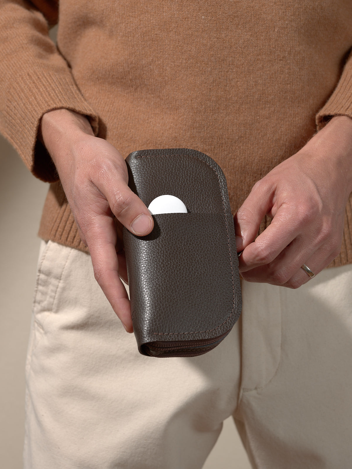 A person's hands holding the closed, dark brown leather glasses case and placing a white, circular object (like a tracker or earbud case) into a small exterior slip pocket.