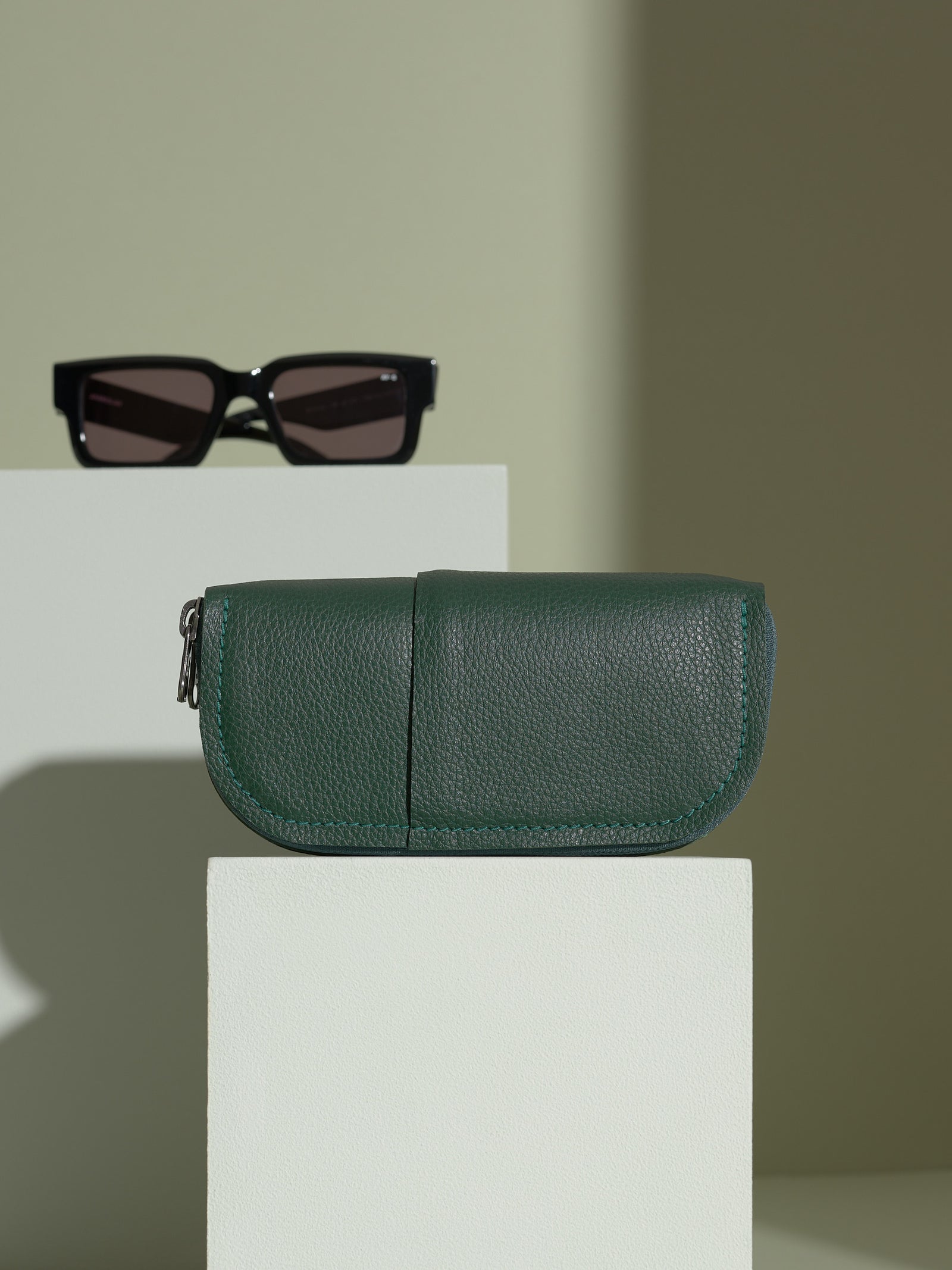 The closed, green Capra leather double glasses case resting horizontally on two white platforms, with a pair of black rectangular sunglasses positioned behind it on the upper platform.