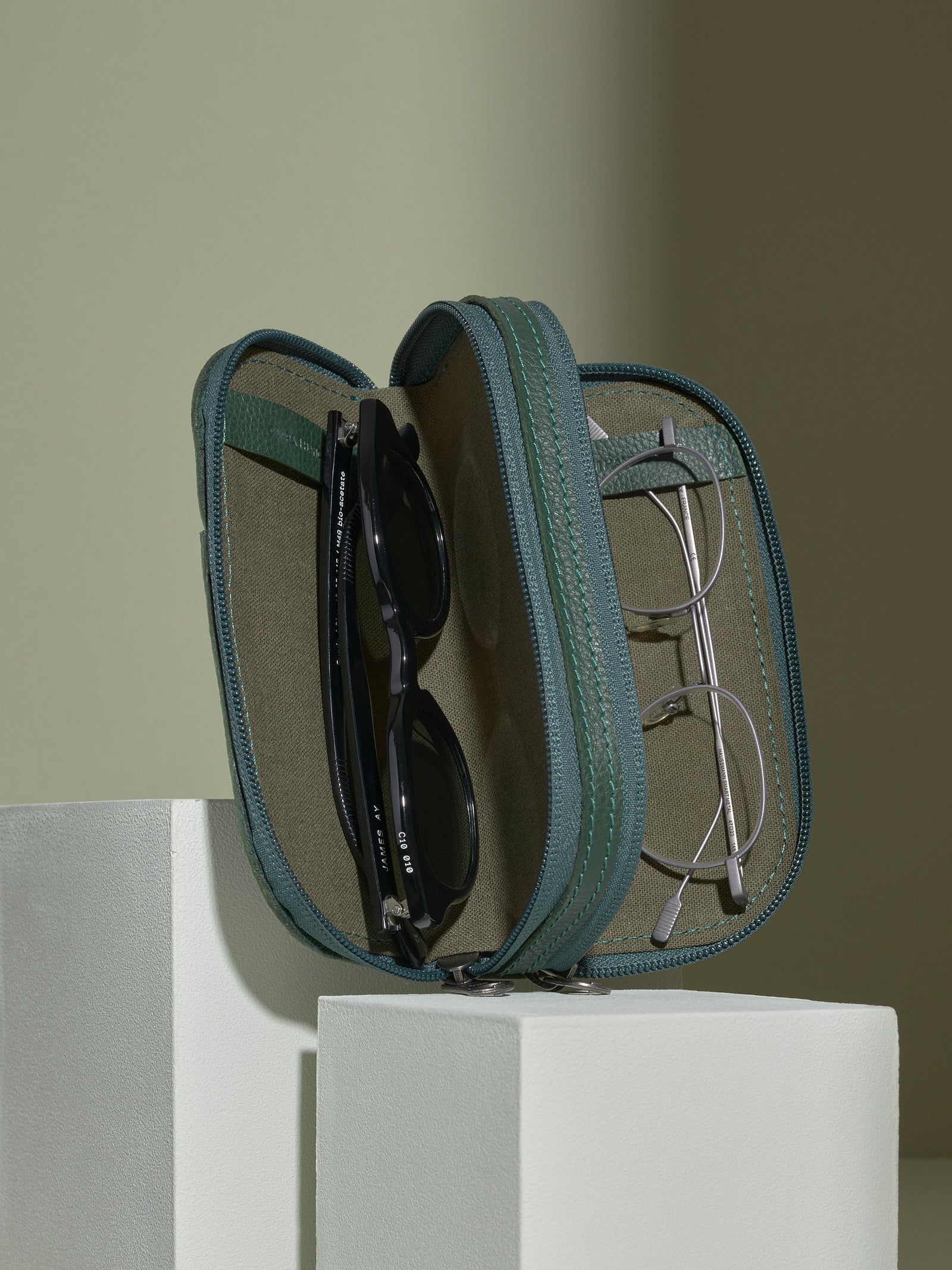 The opened, green leather double glasses case standing upright on white platforms, displaying two pairs of glasses stored inside: black sunglasses on the left and round silver-framed clear glasses on the right.