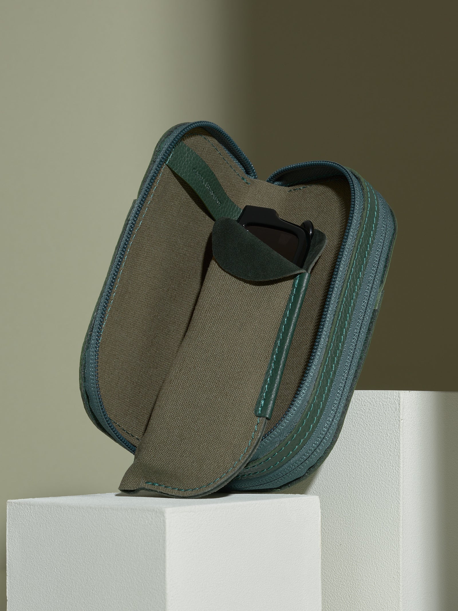 The opened, zipped green leather glasses case standing upright on white platforms, showing one soft olive-green fabric sleeve partially pulled out and containing a pair of black sunglasses.