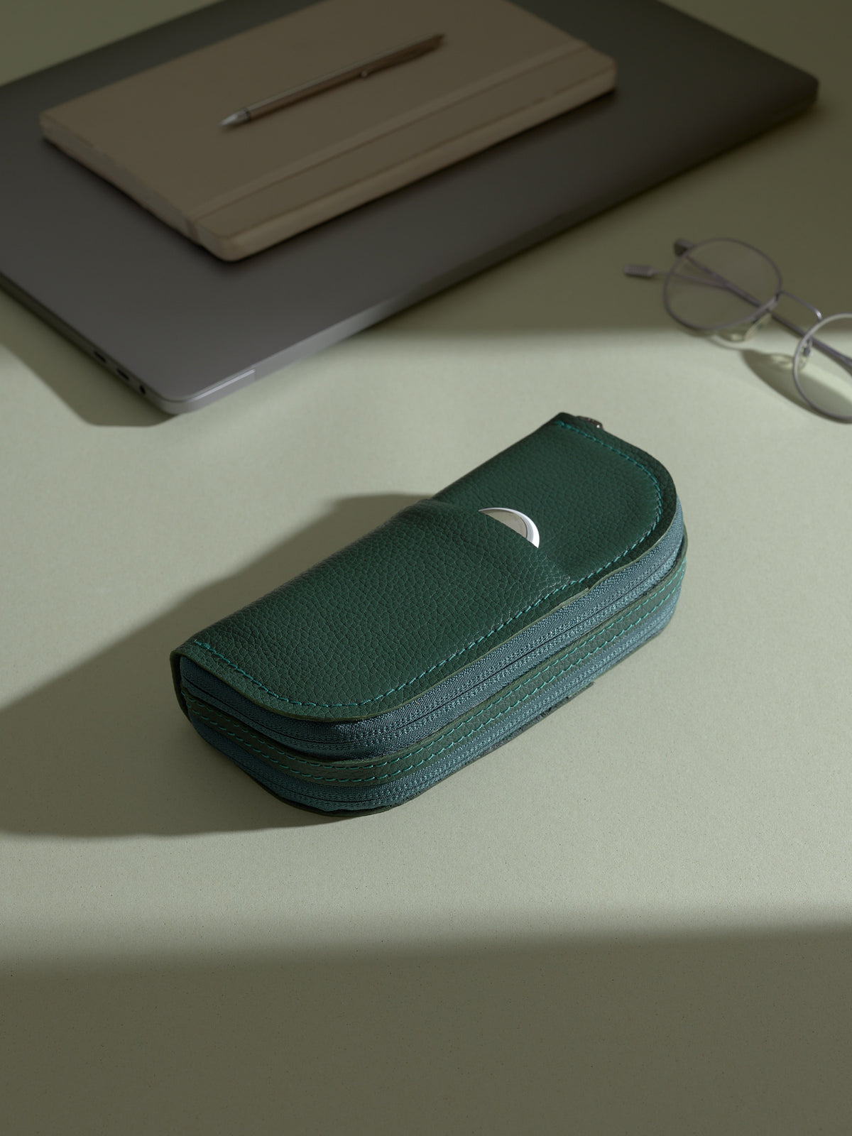 The closed, green Capra leather double glasses case resting on a light green desk, with a white circular object (likely an AirTag or earbud case) partially visible in the exterior slip pocket. A laptop, notepad, and glasses are in the background.